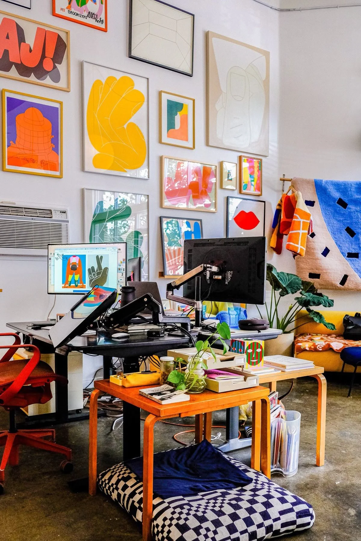 A colorful artist's workspace with multiple computer monitors, framed abstract art on the wall, a small wooden table with plants, books, and art supplies, a cushioned floor mat, and a yellow sofa with patterned textiles.