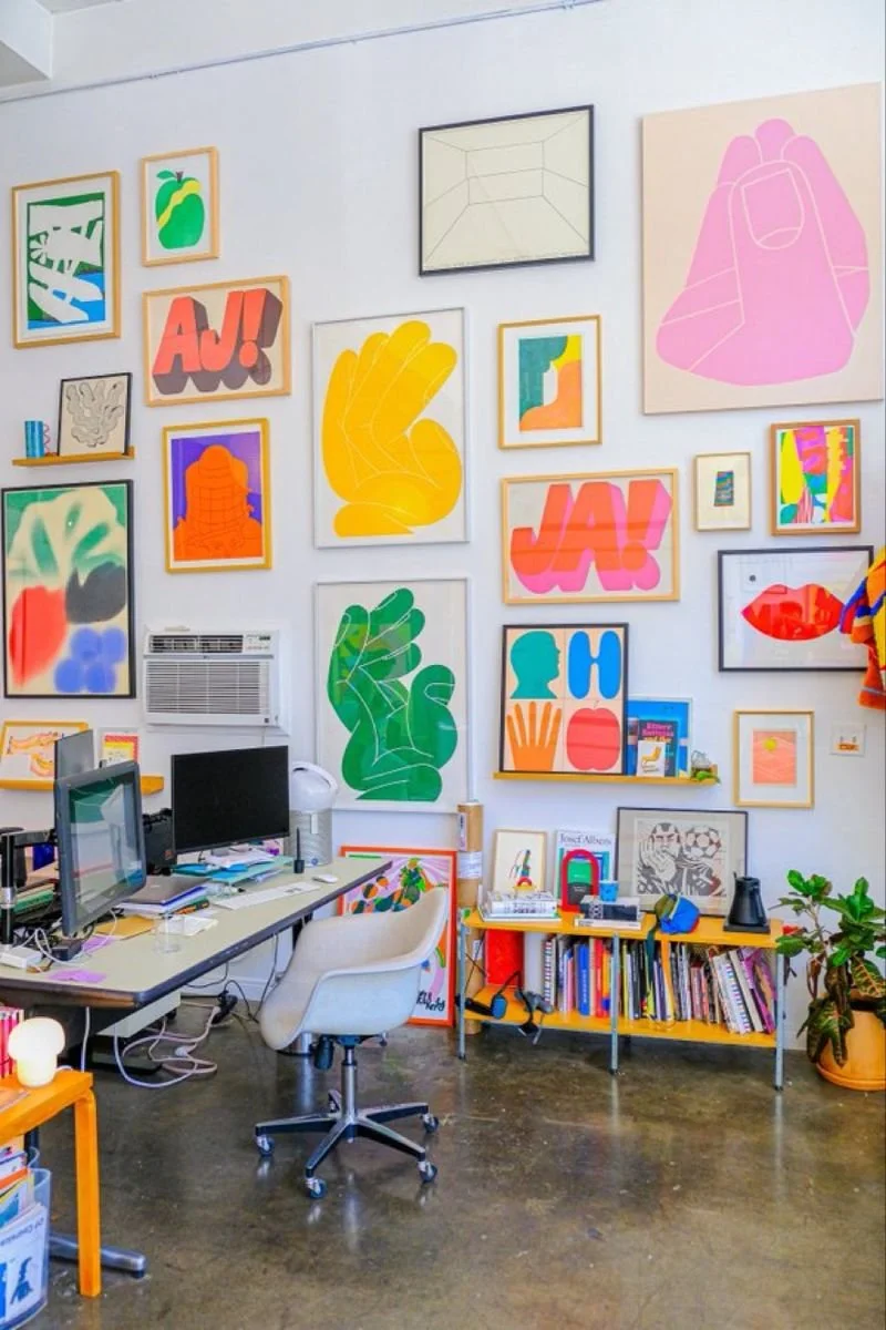 Colorful art gallery wall with various framed and unframed abstract and graphic art prints, including hand shapes, lips, and text, with a desk, computer, books, and potted plant in the foreground.
