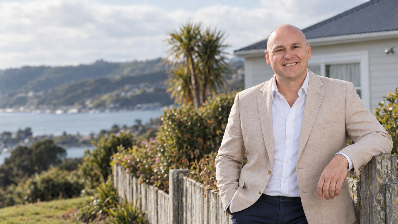 aBlair Thompson, Porirua real estate agent, leaning on a picket fence outside a weatherboard home with harbour views