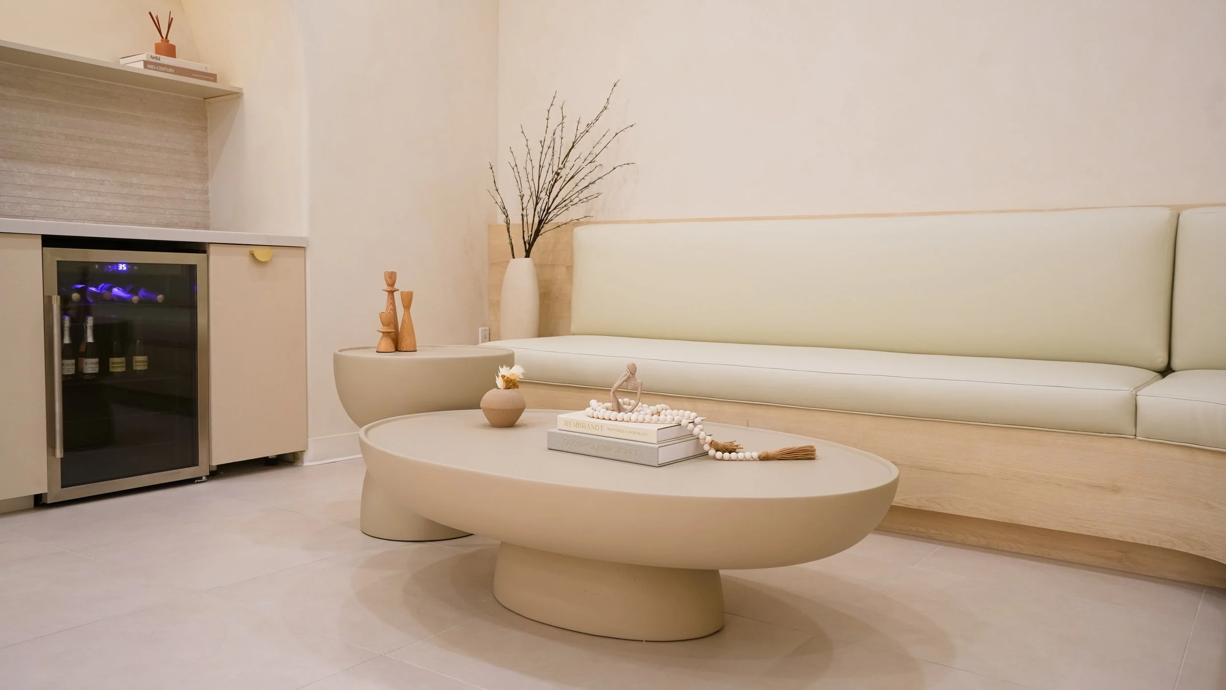 Minimalist living room with beige sofa, oval coffee table with decorative items, tall vase with branches, and a mini refrigerator in the corner.