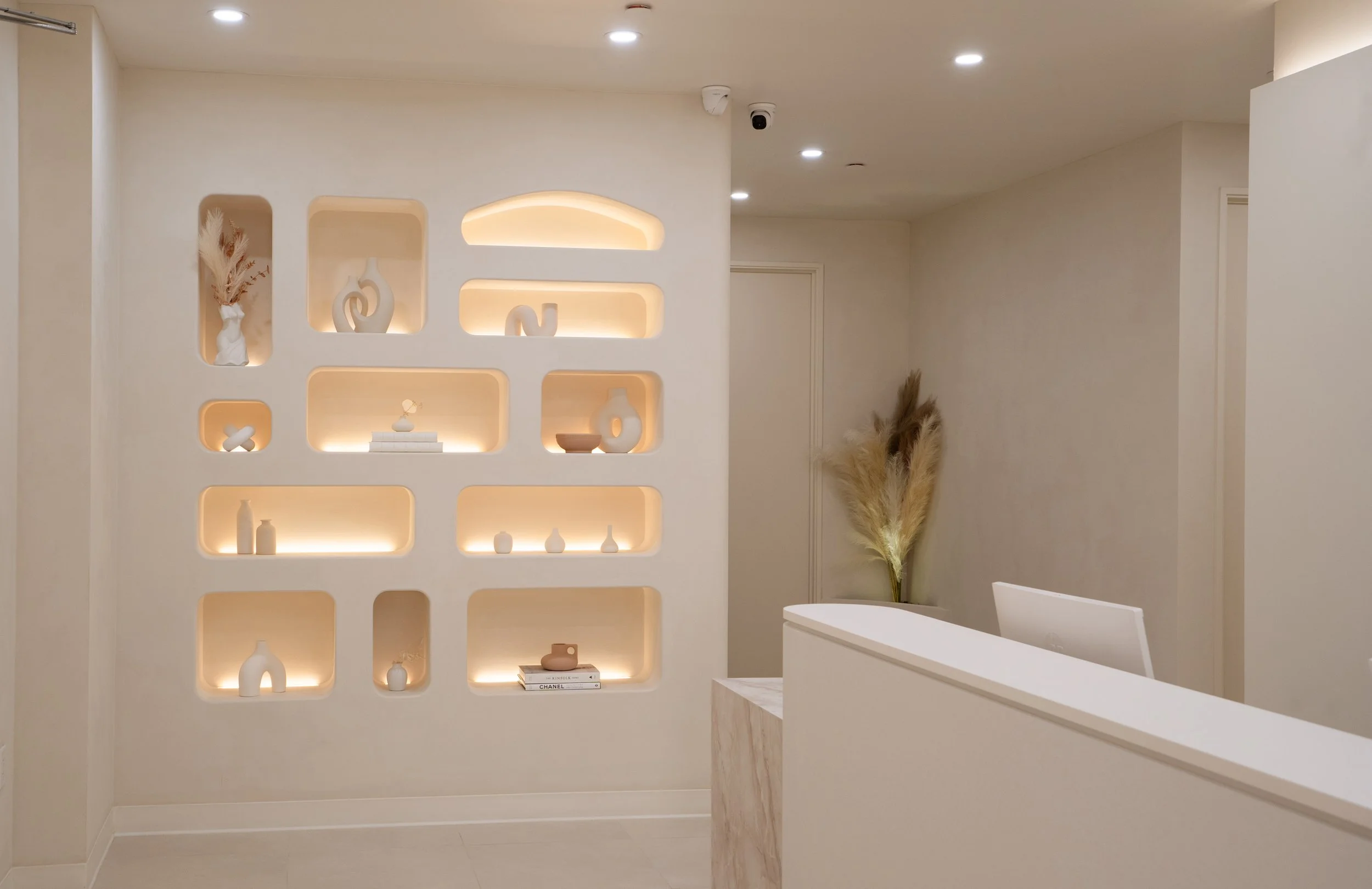 Modern interior with built-in wall shelves displaying decorative vases and books, illuminated by ambient lighting, with a reception desk and pampas grass decor in the corner.