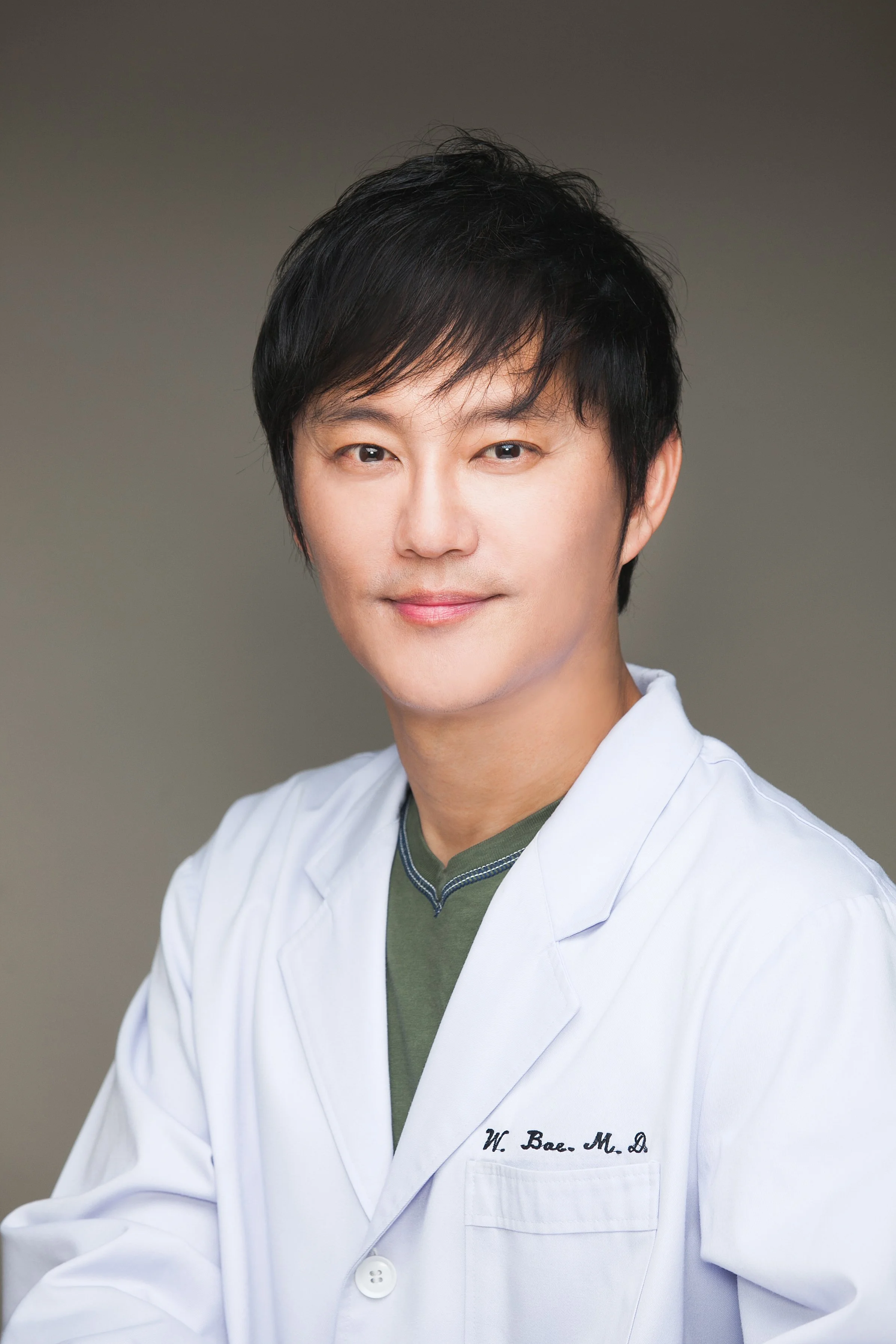 A portrait of a man with black hair wearing a white lab coat and green shirt, smiling softly against a neutral background.