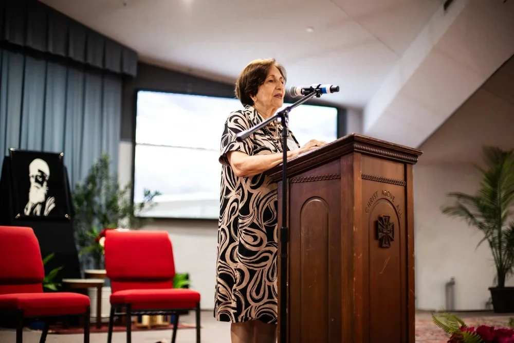 A woman in a white sleeveless top and orange floral skirt speaks into a microphone on a stage next to a wooden pulpit with a decorative cloth. Photo from past Florida Southern Flame Baha'i Summer School