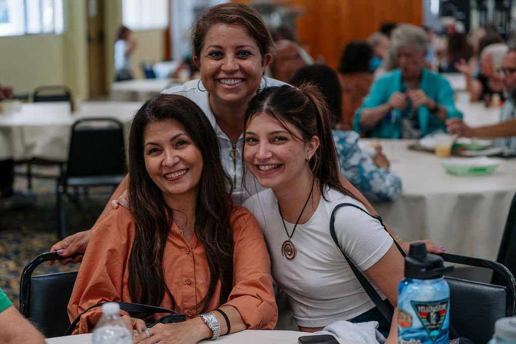 Three women smiling at a social gathering, sitting at a table with people in the background.