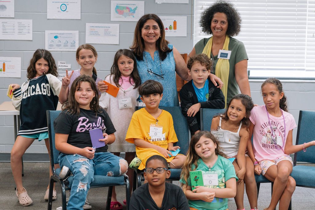 A group of diverse children sitting and standing, smiling at the camera in a classroom. Two adult women stand behind them. Charts and educational posters are on the wall in the background. Children's class at Baha'i Summer school in Florida