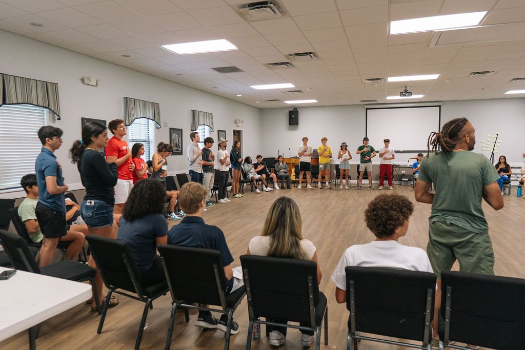 Group of people standing and sitting in a circle in a community room, participating in an interactive group activity. Youth activities at Southern Flame Baha'i Summer School