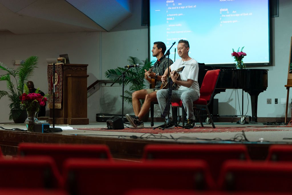 Two men playing guitars on stage in a church setting, with a projector screen displaying lyrics in the background.
