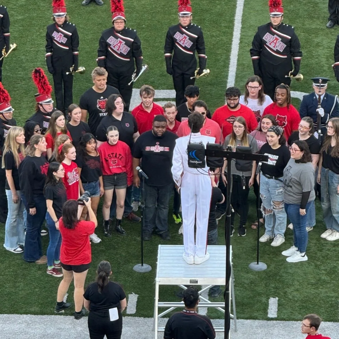 A group of students and band members gathered on a football field to participate in a ceremony, with one person standing on a platform in front of microphones, facing the crowd.