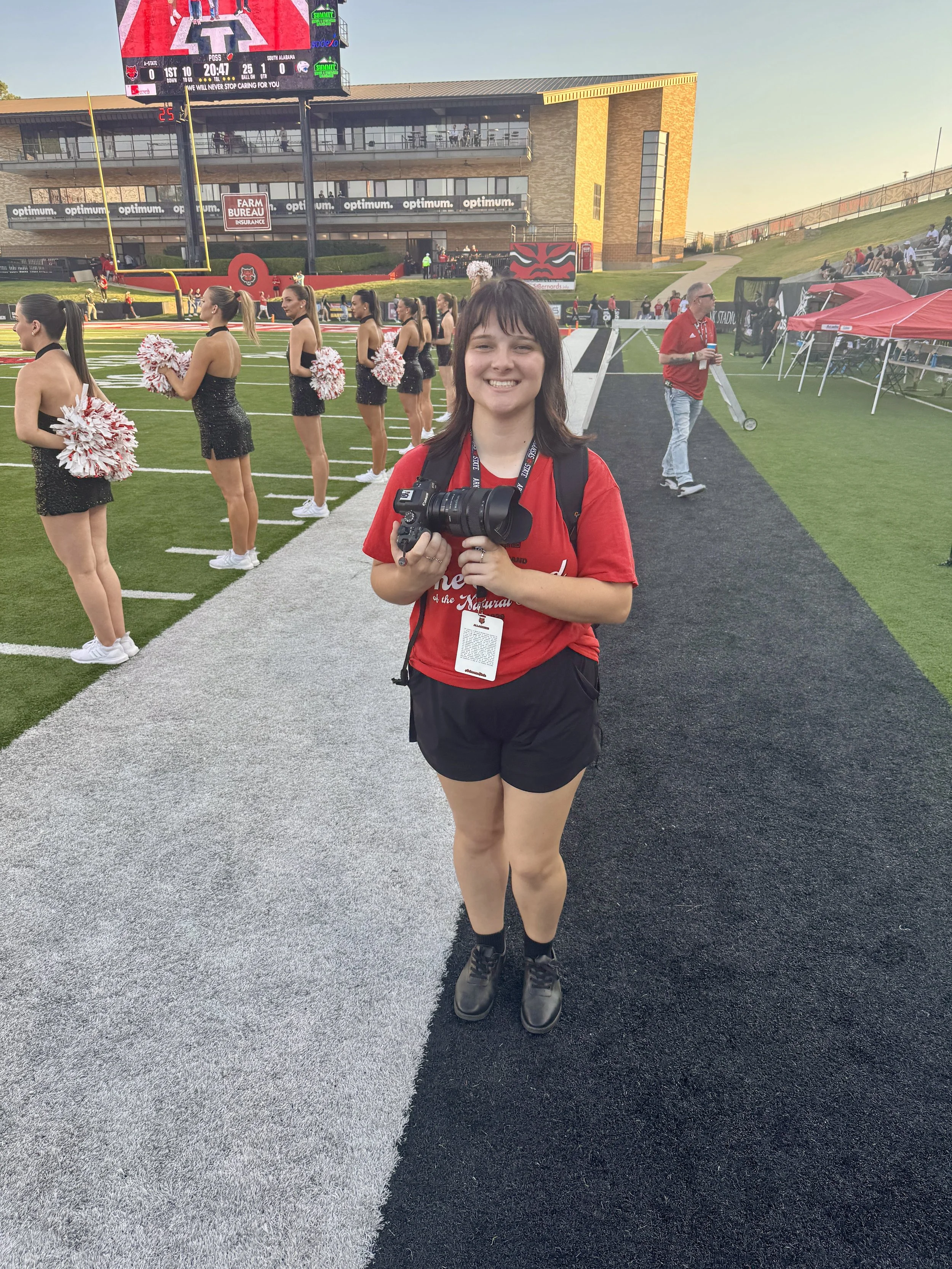 A young woman holding a camera and smiling at the camera on a football field with cheerleaders lined up behind her, cheerleaders holding pom-poms, and a large scoreboard in the background during a game.