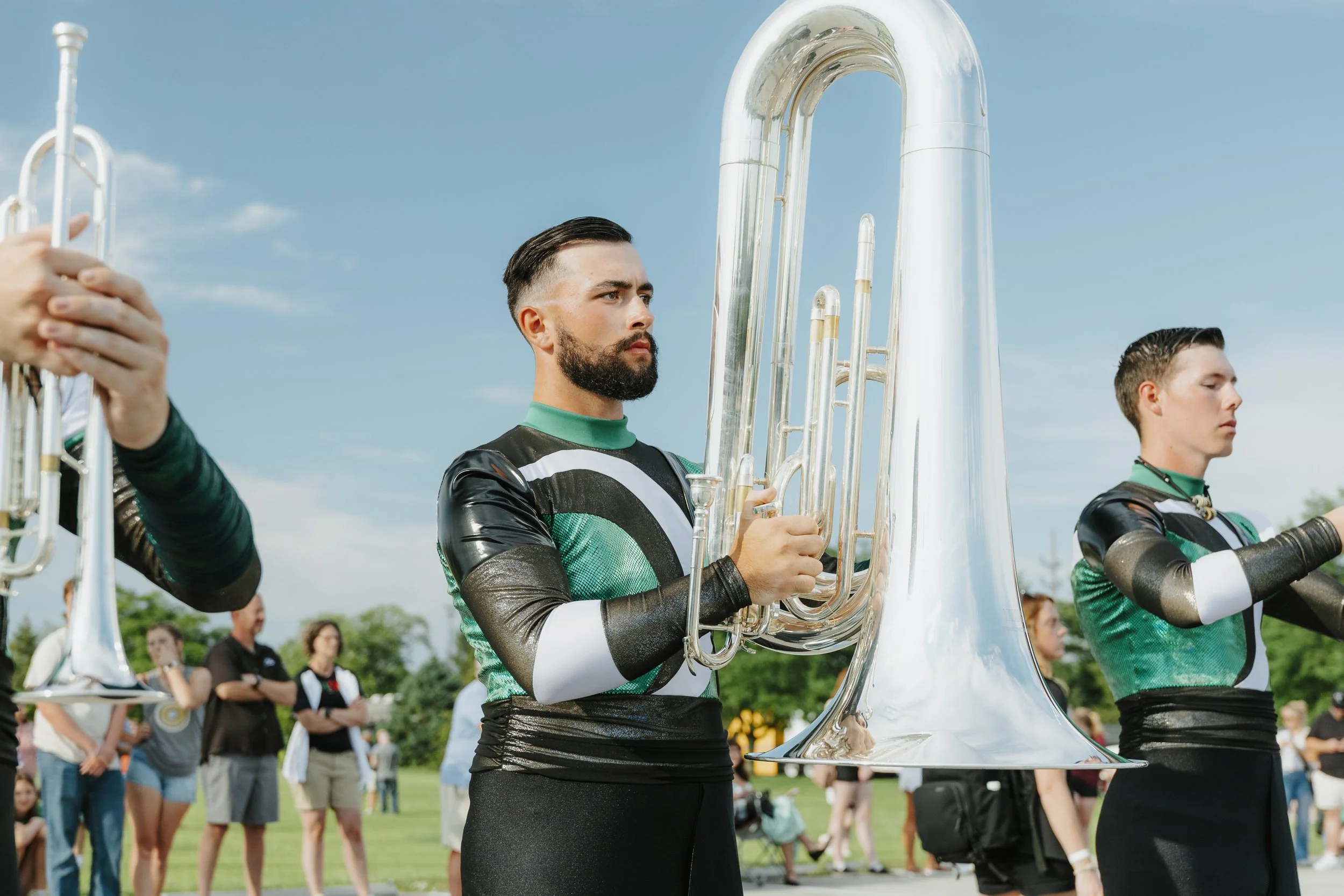 Two band members holding large brass instruments, standing outdoors in a sunny field with an audience in the background.