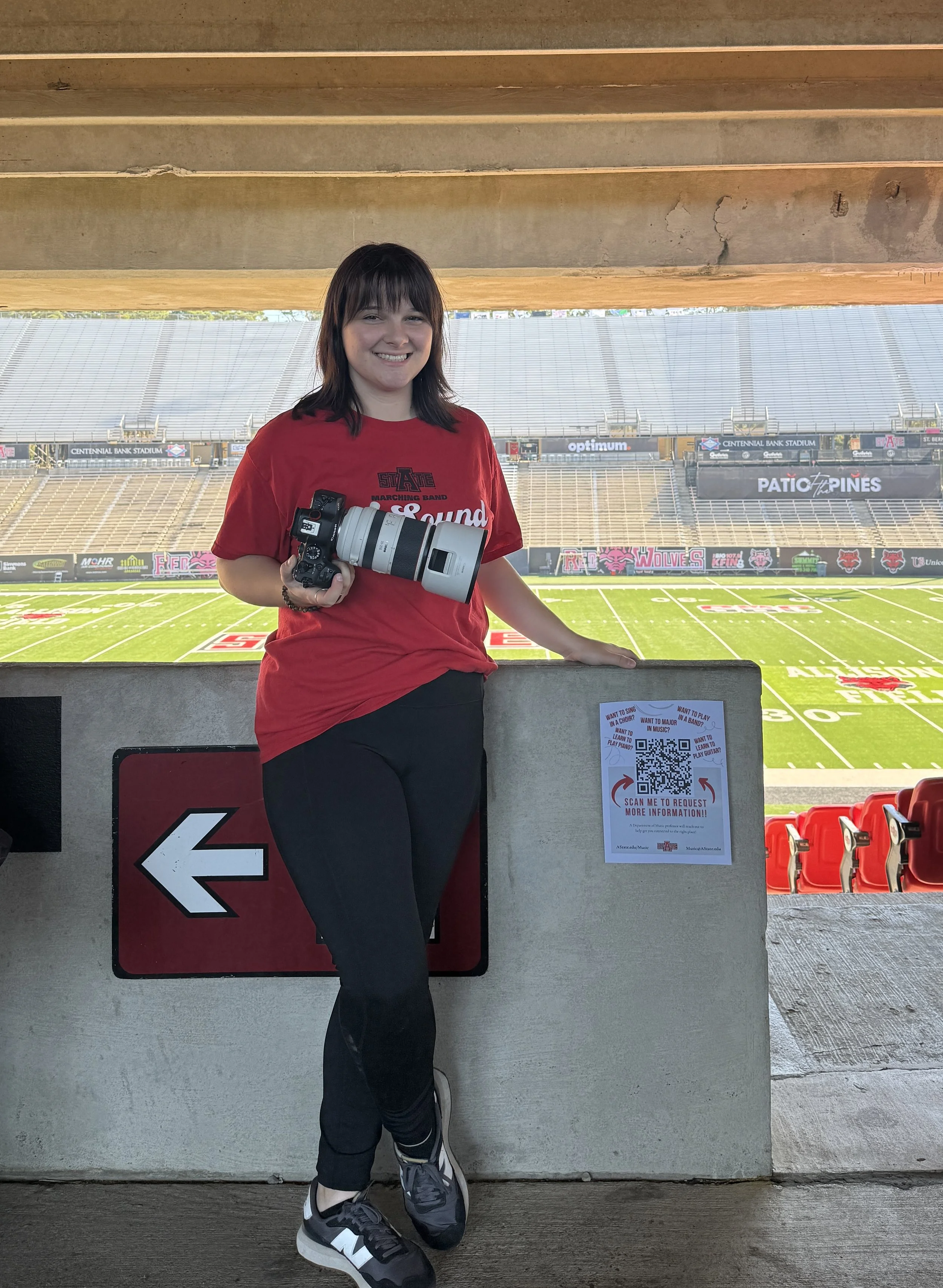 A young woman standing at a stadium with a camera in her hand, smiling. The stadium seats are visible in the background, and there is a QR code sign on a wall beside her.