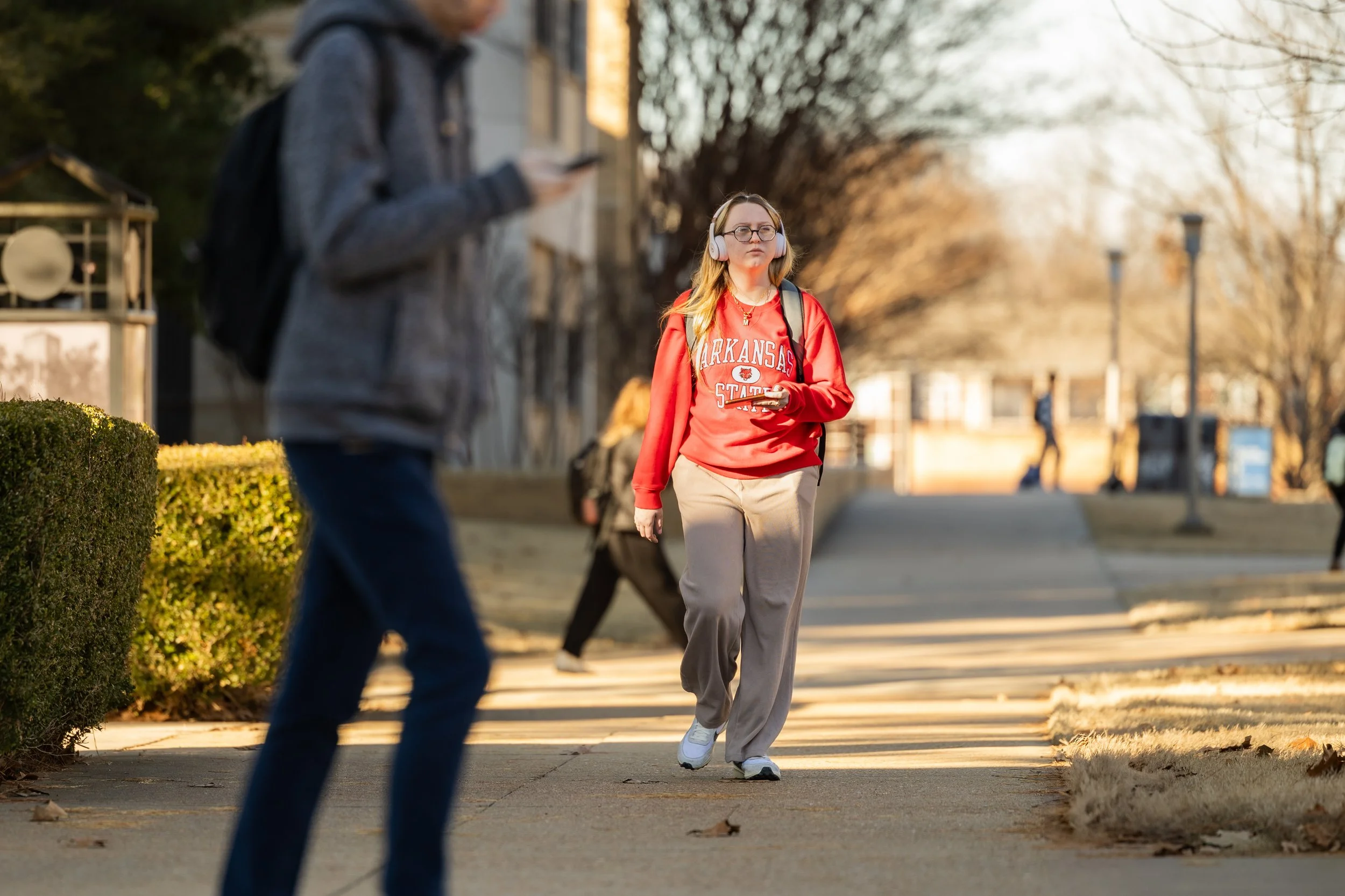A young woman with long blonde hair, glasses, and headphones, wearing a red Arkansas State sweatshirt and beige sweatpants, walks on a sidewalk while looking at her phone. Other pedestrians are seen in the background on a sunny day with leafless tree