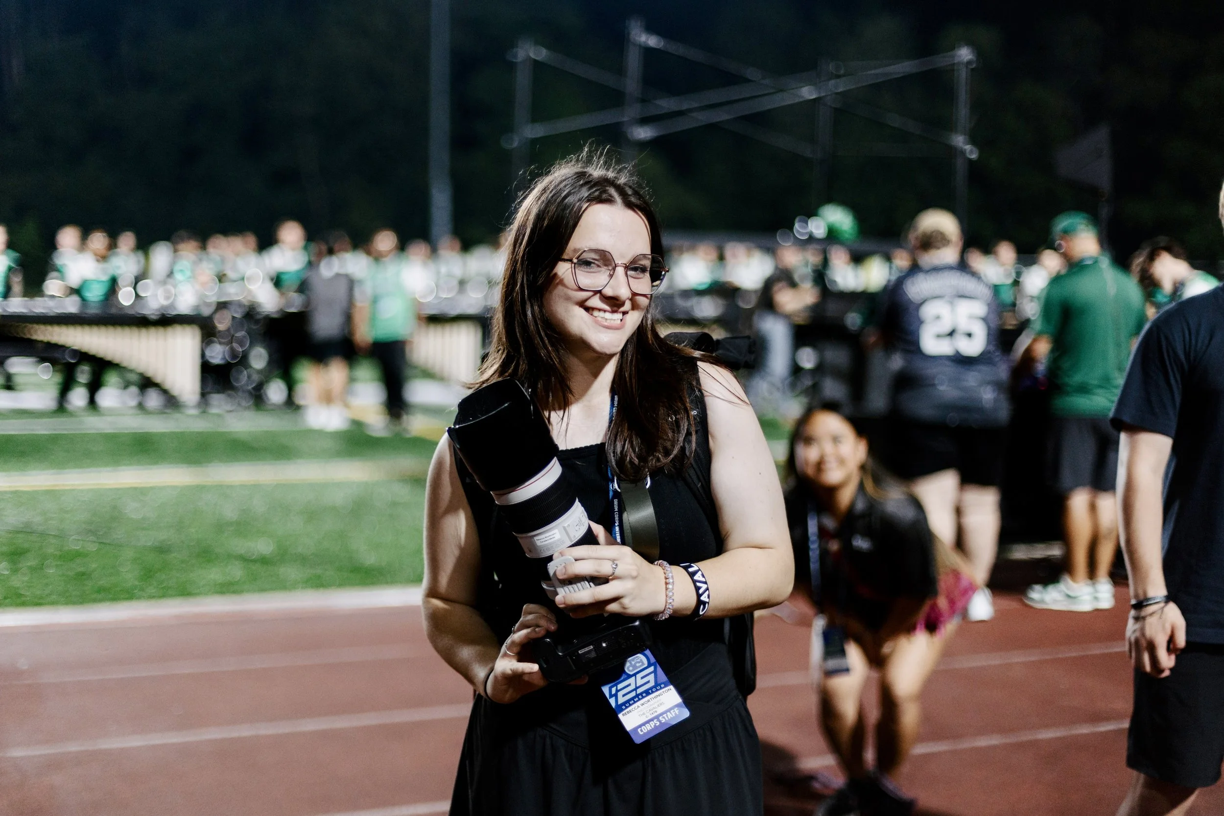 A young woman with glasses and a black sleeveless top smiling, holding a professional camera with a large telephoto lens, at an outdoor event on a sports field at night, with a marching band performing in the background.
