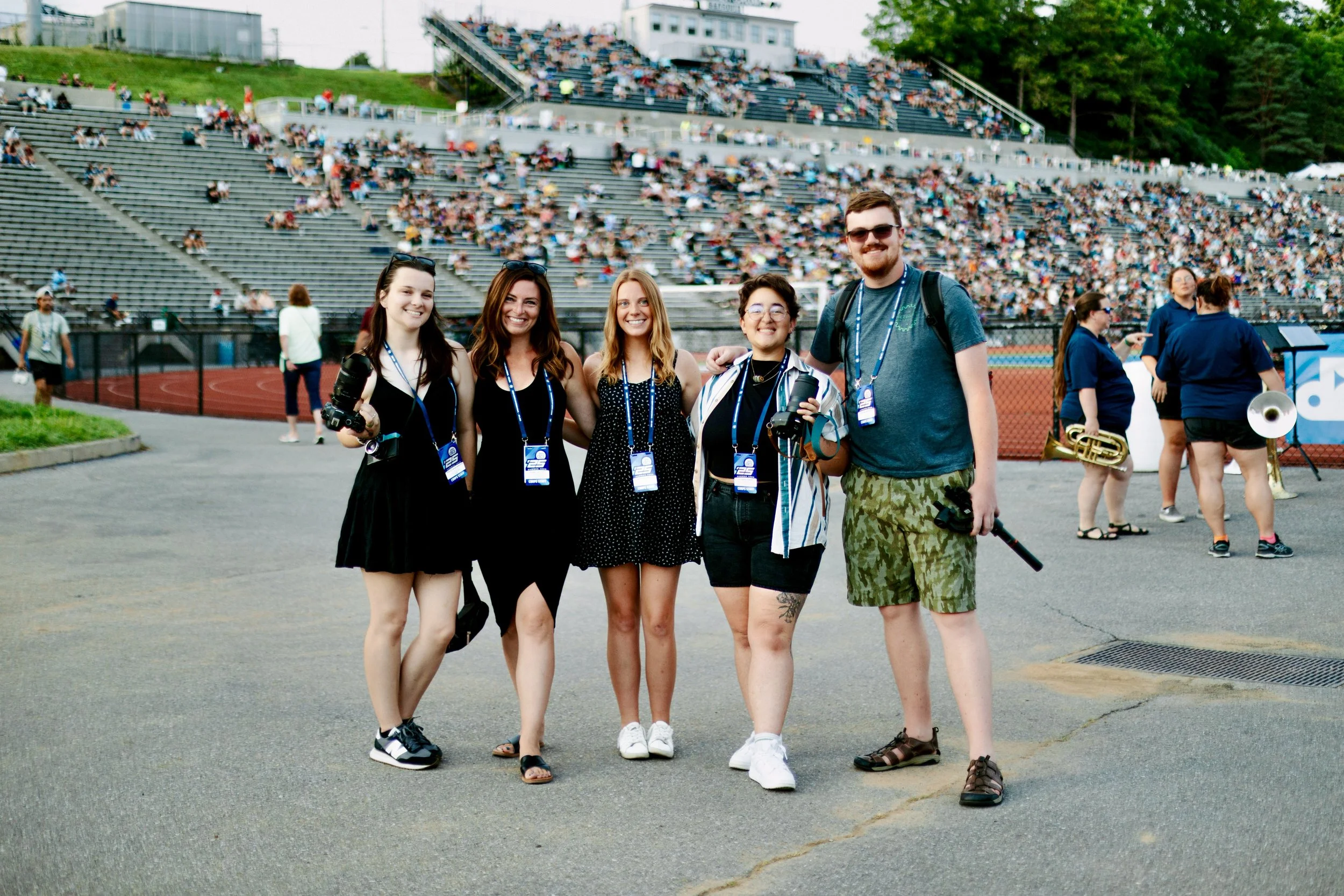 Group of five young adults standing together at a stadium event, smiling, with some holding cameras, surrounded by a crowd and musical band members in the background.