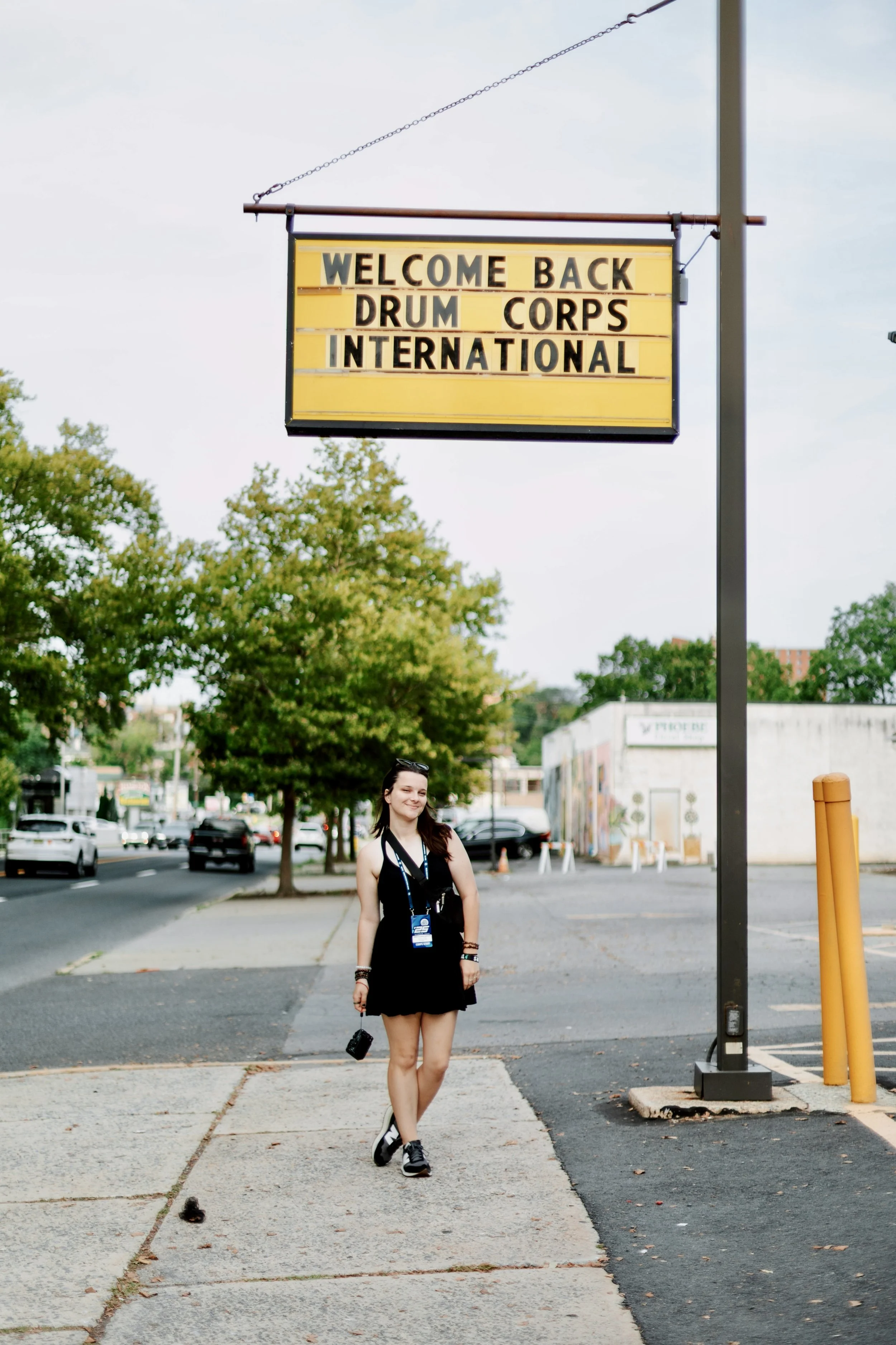 A young woman walking on a sidewalk near a street, with a welcome sign hanging above her that reads: "Welcome Back Drum Corps International." She is wearing a black dress and sneakers, carrying a camera, and has a badge around her neck.