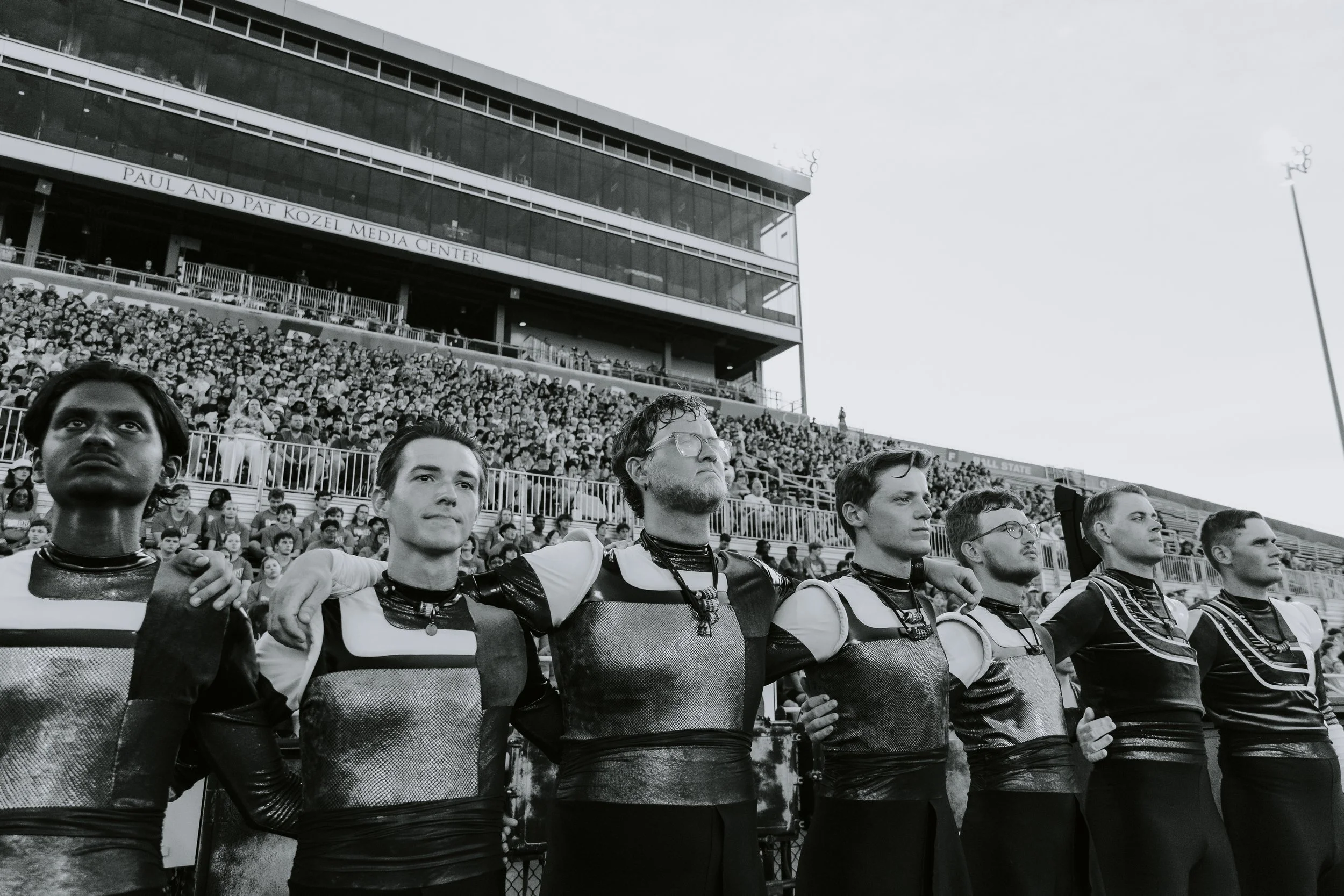 A group of six football players standing arm in arm on the field, with a large crowd in the stadium stands behind them. The players are wearing matching uniforms, and the stadium sign reads "Paul and Pat Kozel Media Center."