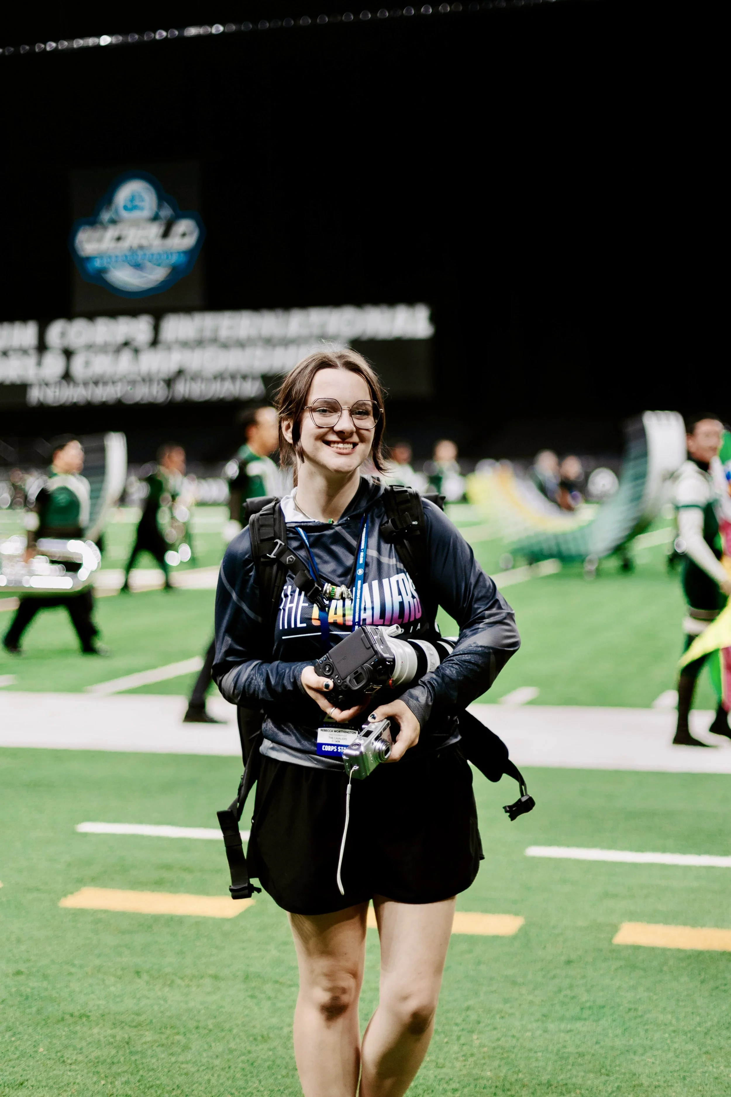 A young woman smiling and holding camera equipment at an indoor stadium with people and flags in the background.