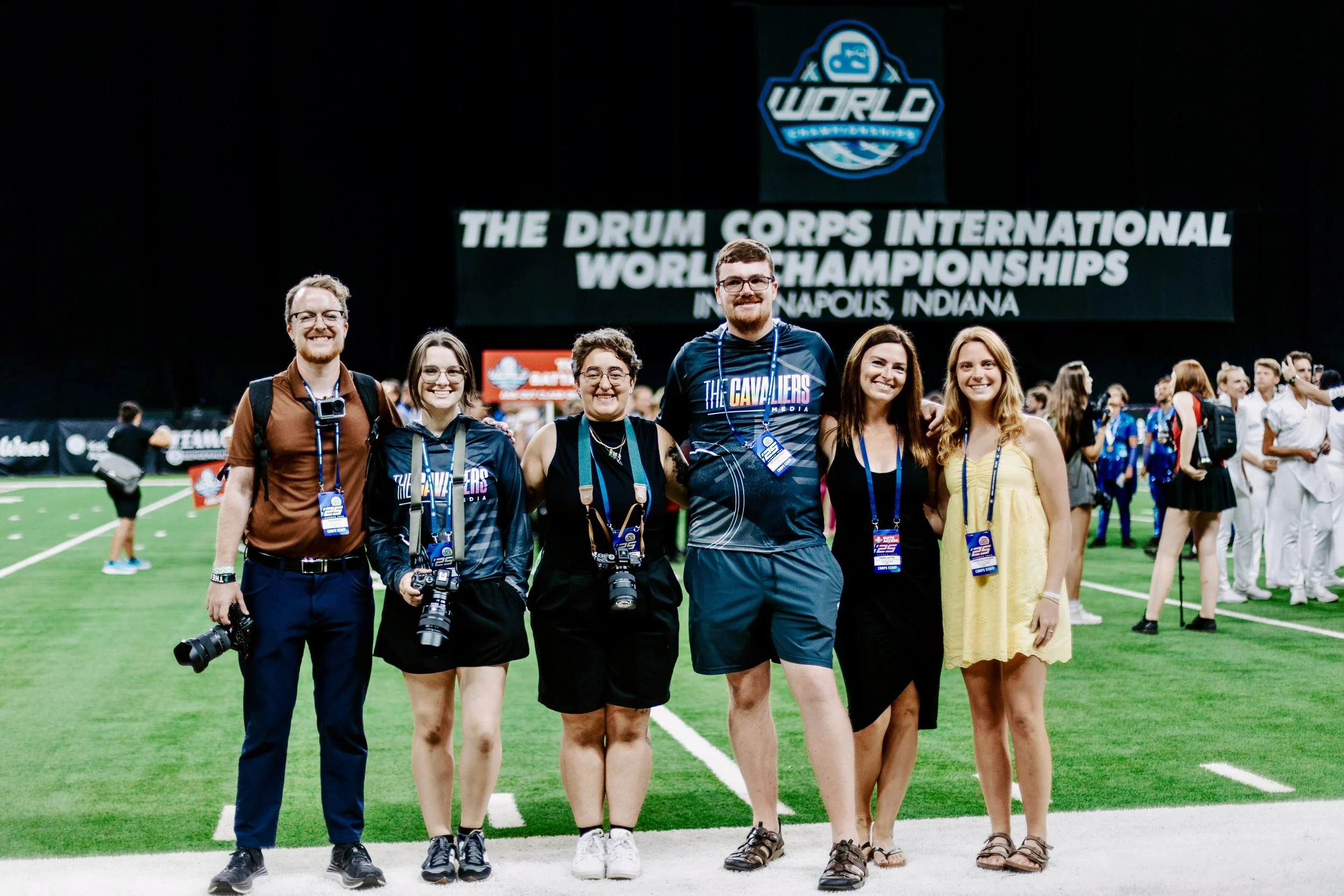Group of six people standing on a green indoor field at the Drum Corps International World Championships in Indianapolis, Indiana, with cameras and event badges, smiling at the camera.