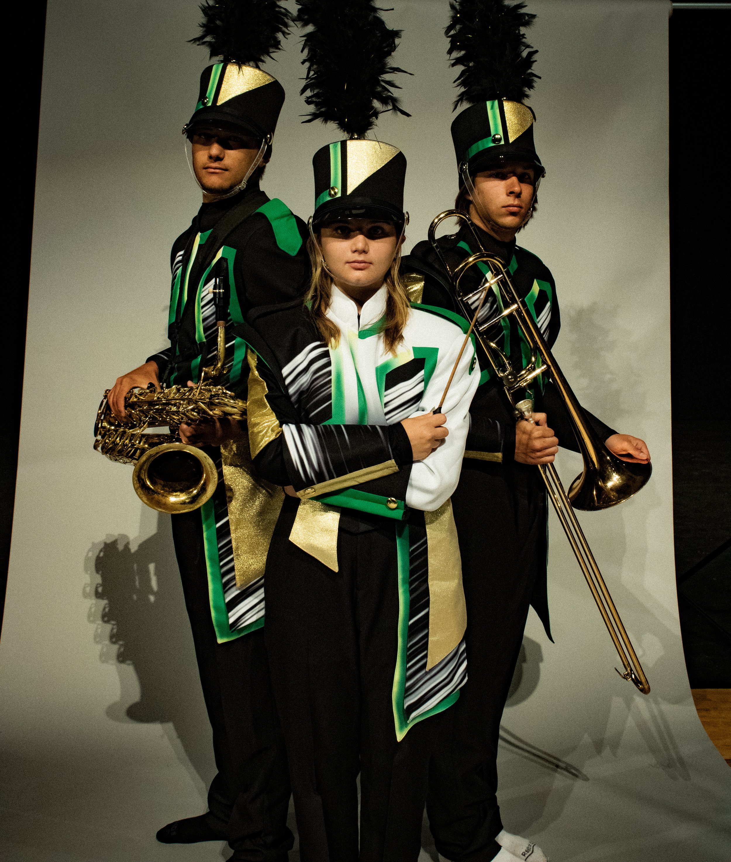 Three young band members dressed in marching band uniforms with black, white, green, and gold colors, holding musical instruments including a saxophone, a trombone, and a clarinet, standing in front of a plain background.