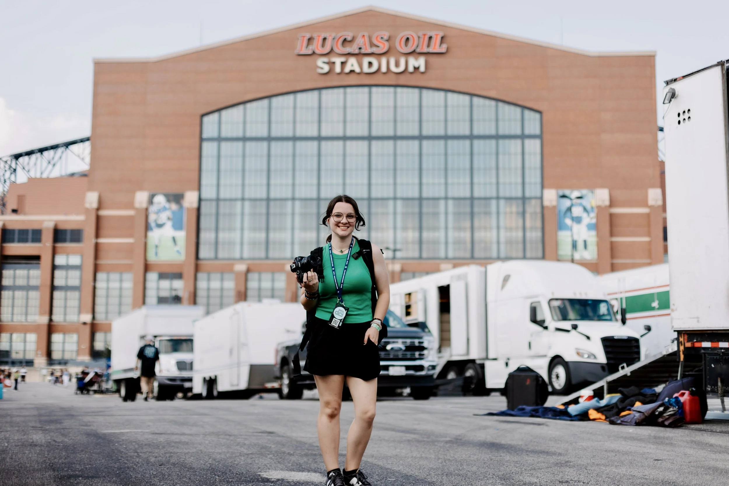 A woman standing outside Lucas Oil Stadium holding a camera, wearing a green top, black shorts, and a backpack, with trucks and people in the background.