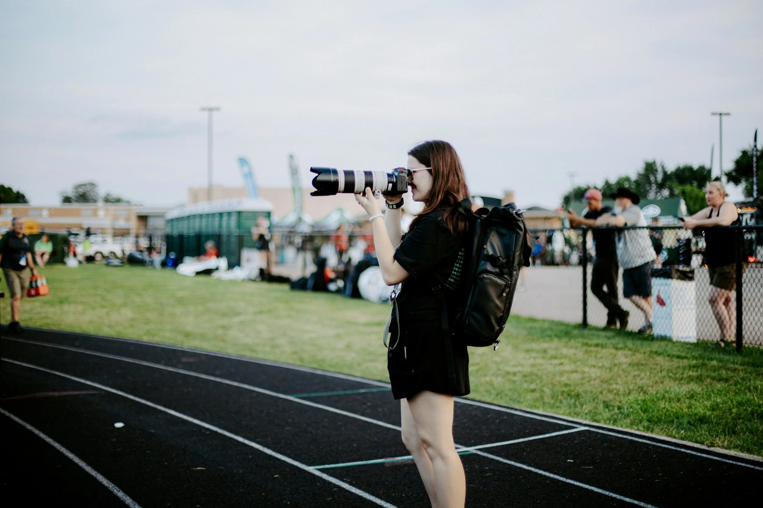 A young woman with brown hair, wearing a black shirt and shorts, stands on a running track, taking photos with a large professional camera with a telephoto lens. She has a black backpack and is facing sideways. In the background, there are people beh
