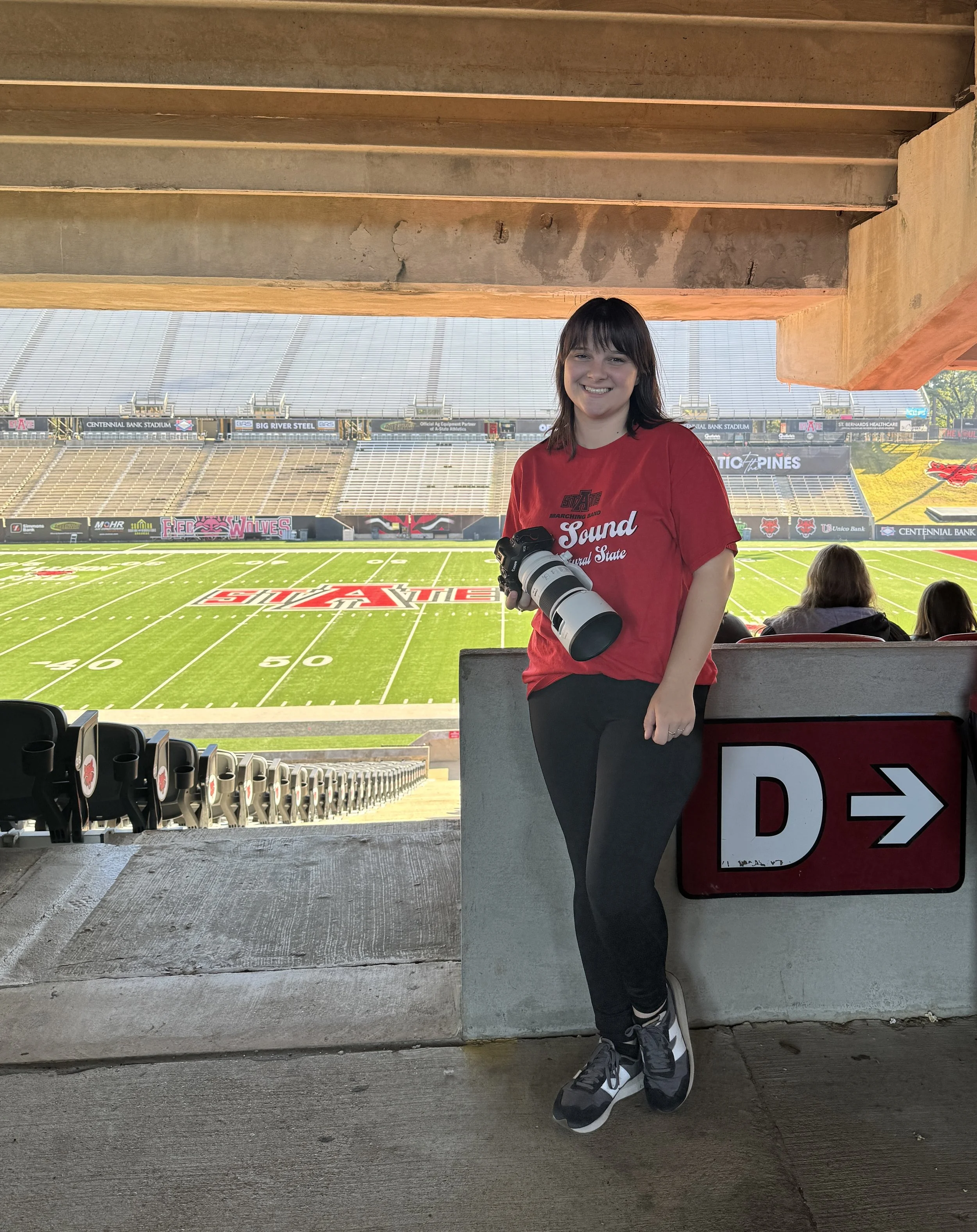 Young woman standing in a stadium seat area with a large camera lens, smiling, holding the camera. She is wearing a red T-shirt and black pants, with a football field in the background.