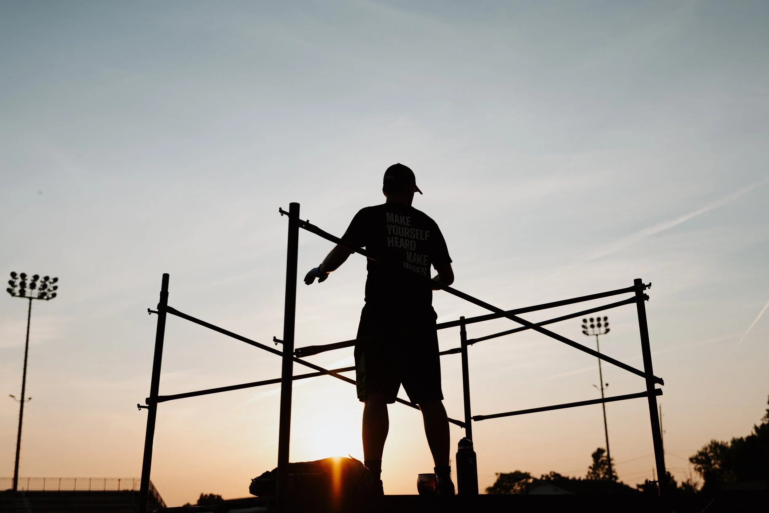 Silhouette of a person working on scaffolding during sunset at an outdoor sports field.