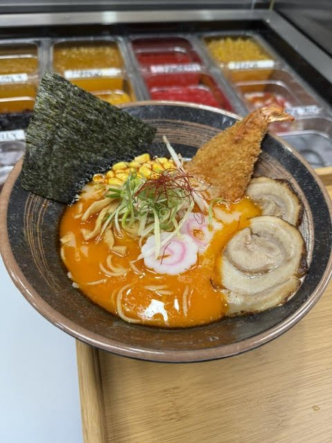 Bowl of ramen with pork slices, fried shrimp, corn, seaweed, and narutomaki, topped with fresh vegetables in a rich broth.