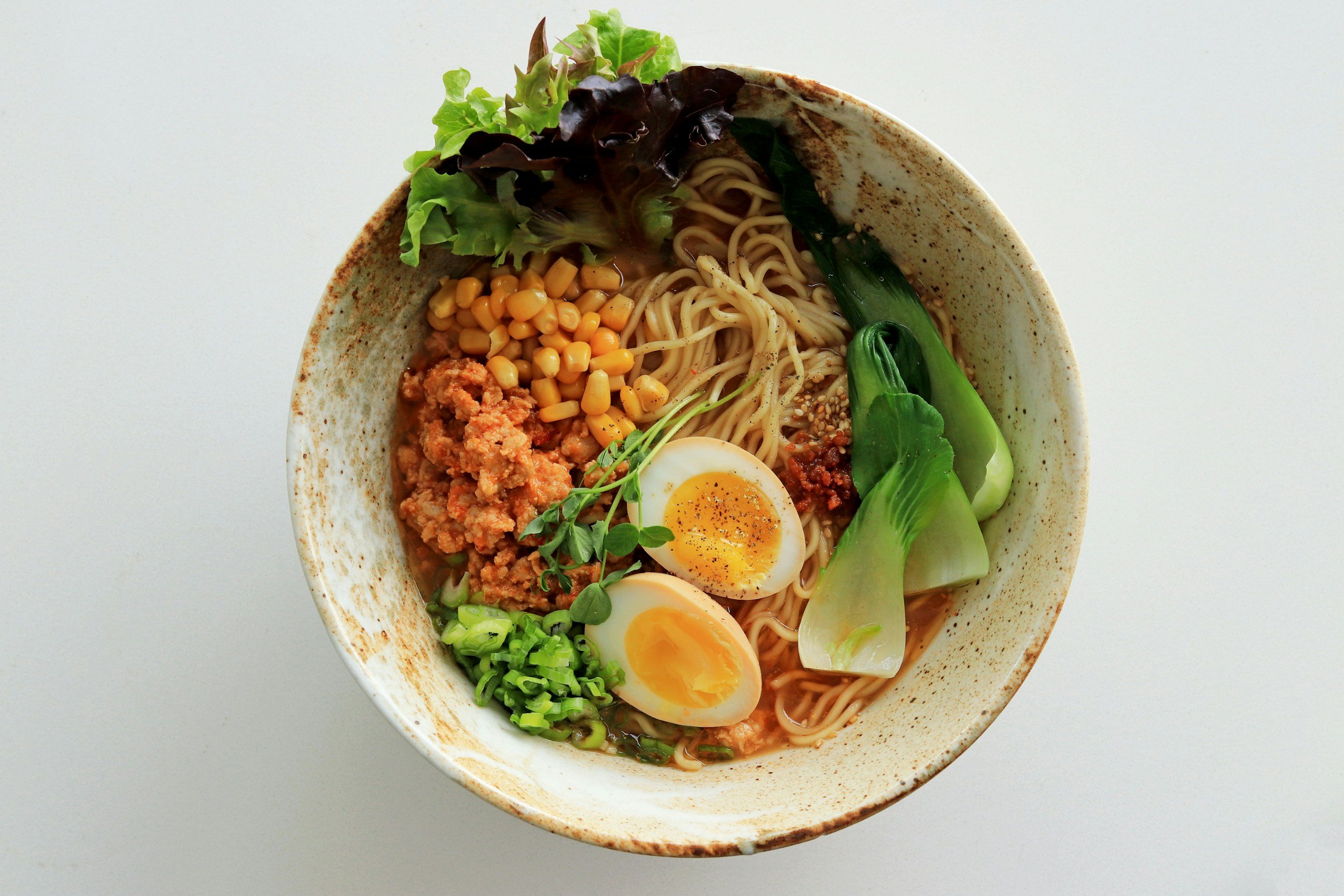 Bowl of ramen with noodles, boiled egg, corn, minced meat, bok choy, and greens.