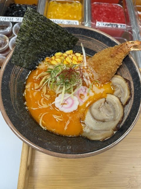Bowl of Japanese ramen with orange broth, topped with sliced pork, shrimp, corn, narutomaki, green onions, and seaweed.