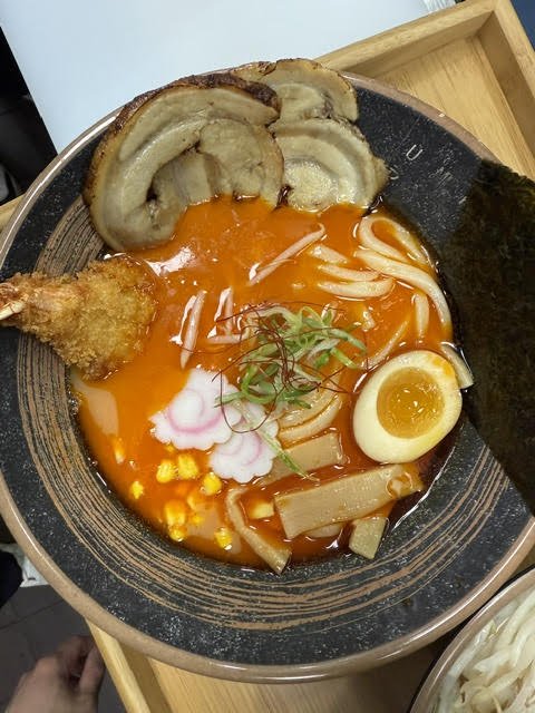 A bowl of spicy ramen with thick broth, featuring sliced pork, a boiled egg, tempura shrimp, fish cake, corn, bamboo shoots, seaweed, and green onions.