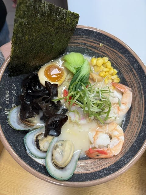 Bowl of ramen with shrimp, clams, egg, seaweed, corn, bok choy, mushrooms, and broth.