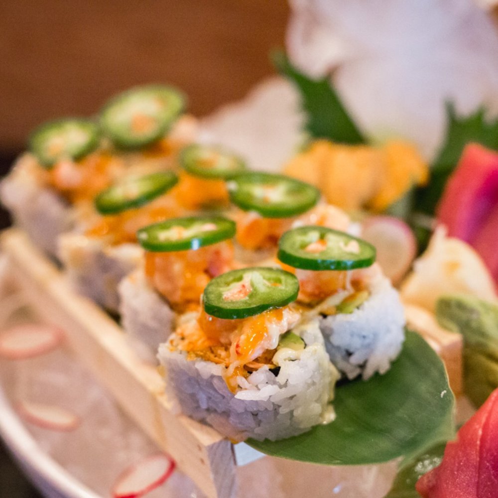 Close-up of sushi rolls topped with jalapeño slices and spicy sauce, placed on a wooden stand.