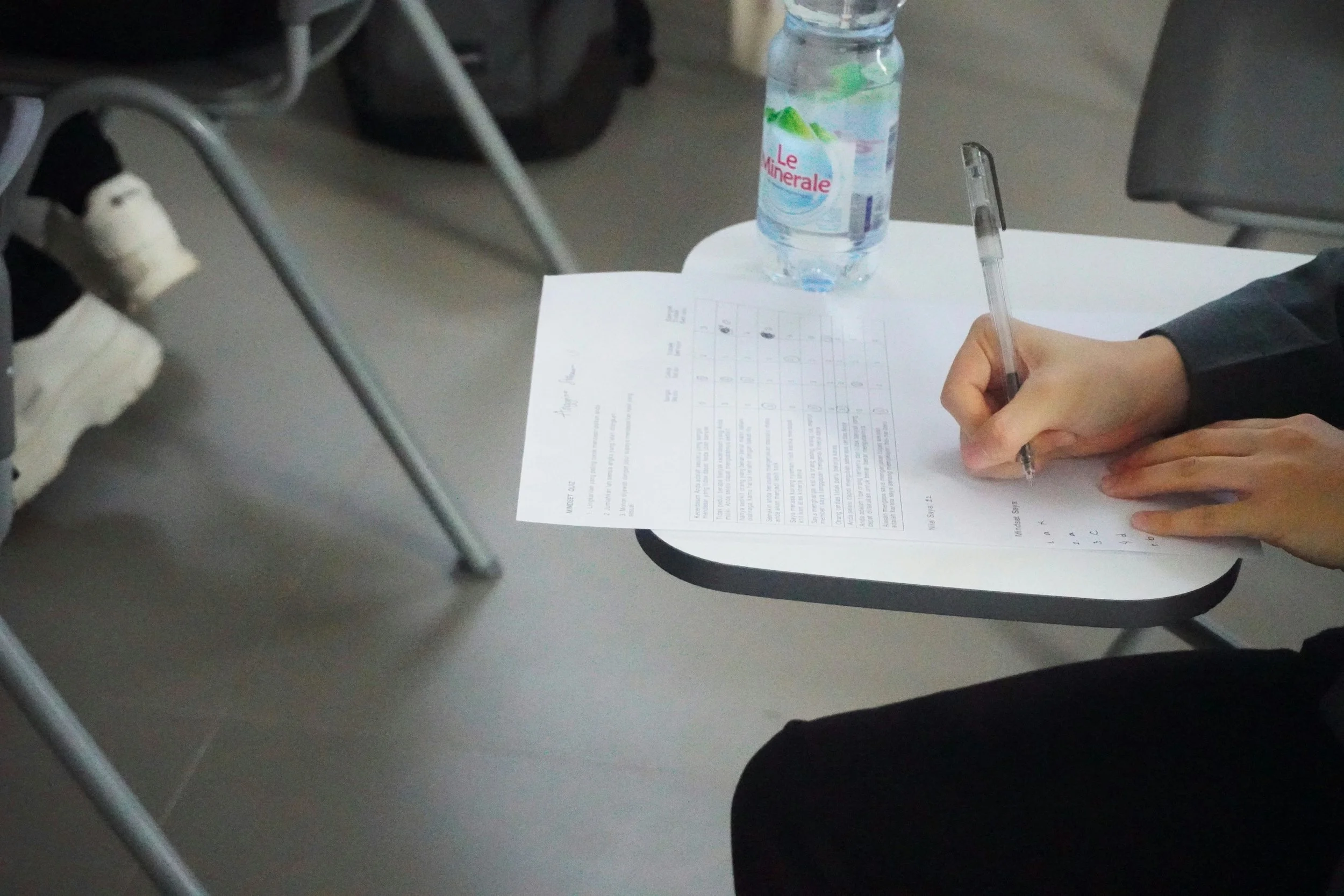 Person's hand writing on a test paper on a desk, with a water bottle nearby and chairs around.