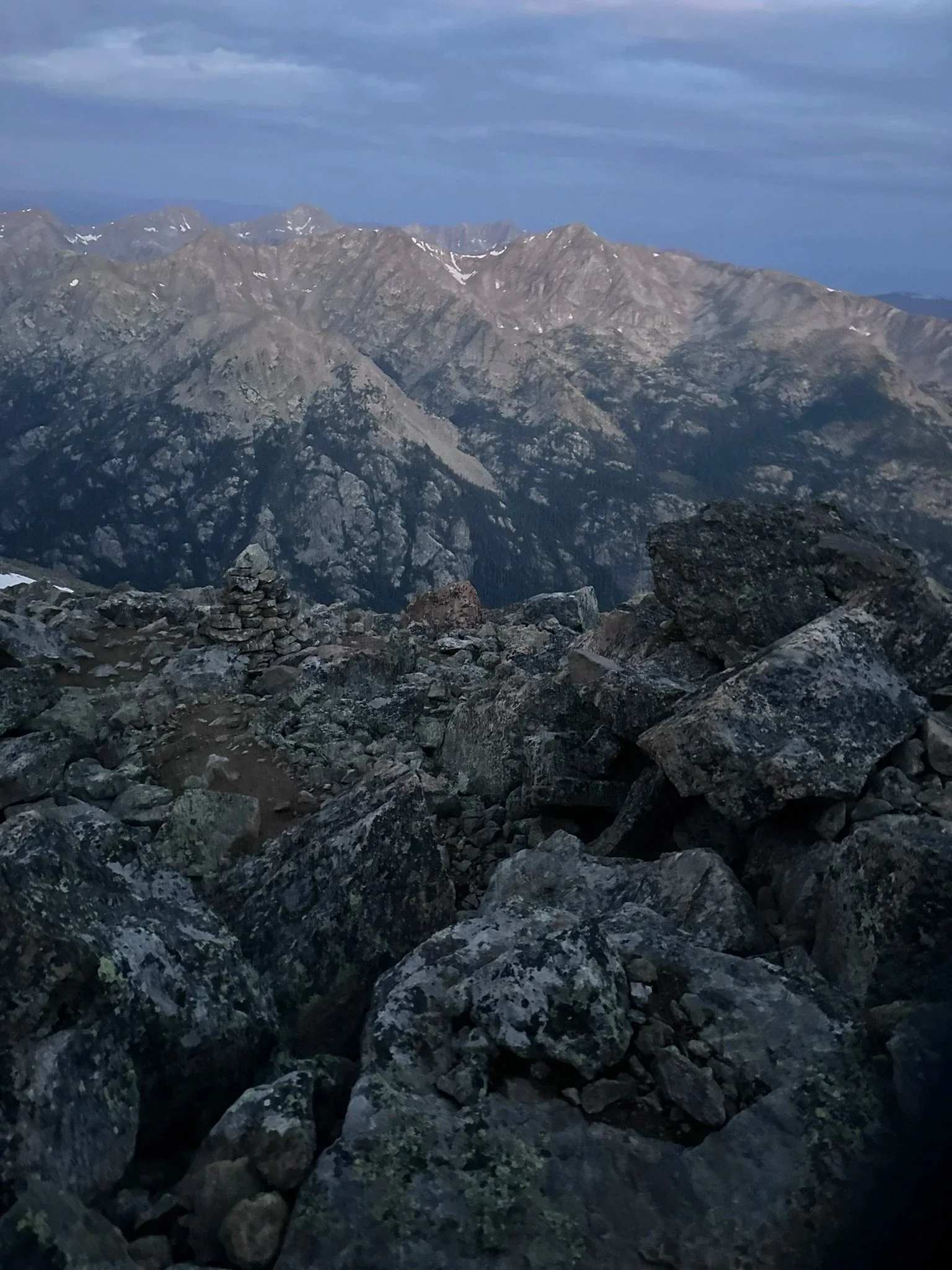 Mountain landscape with rocky foreground and rugged mountain peaks in the background under a cloudy sky.