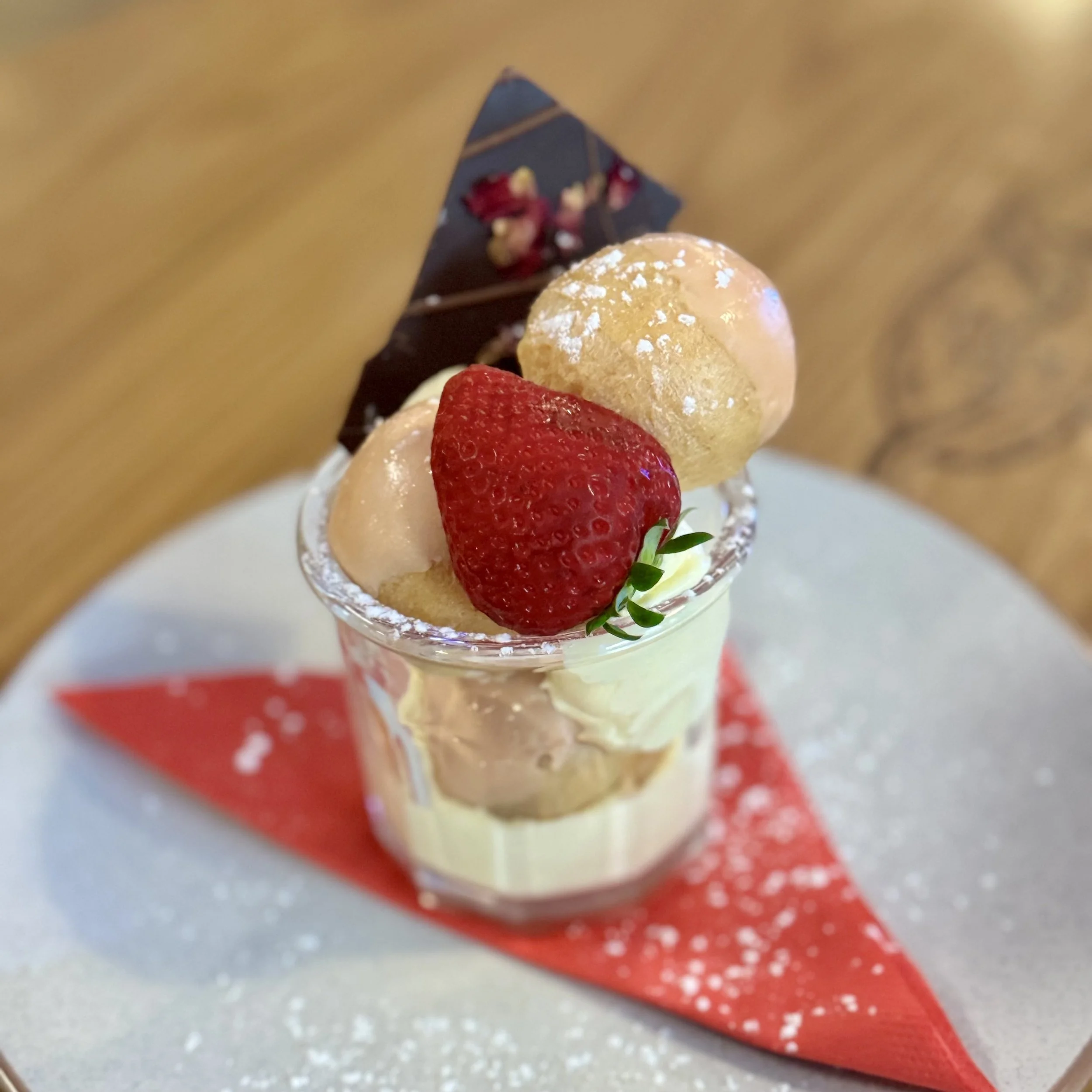 A dessert in a tall glass filled with chopped strawberries and topped with a scoop of vanilla ice cream, placed on a red plate with a red napkin on a wooden table near a window.