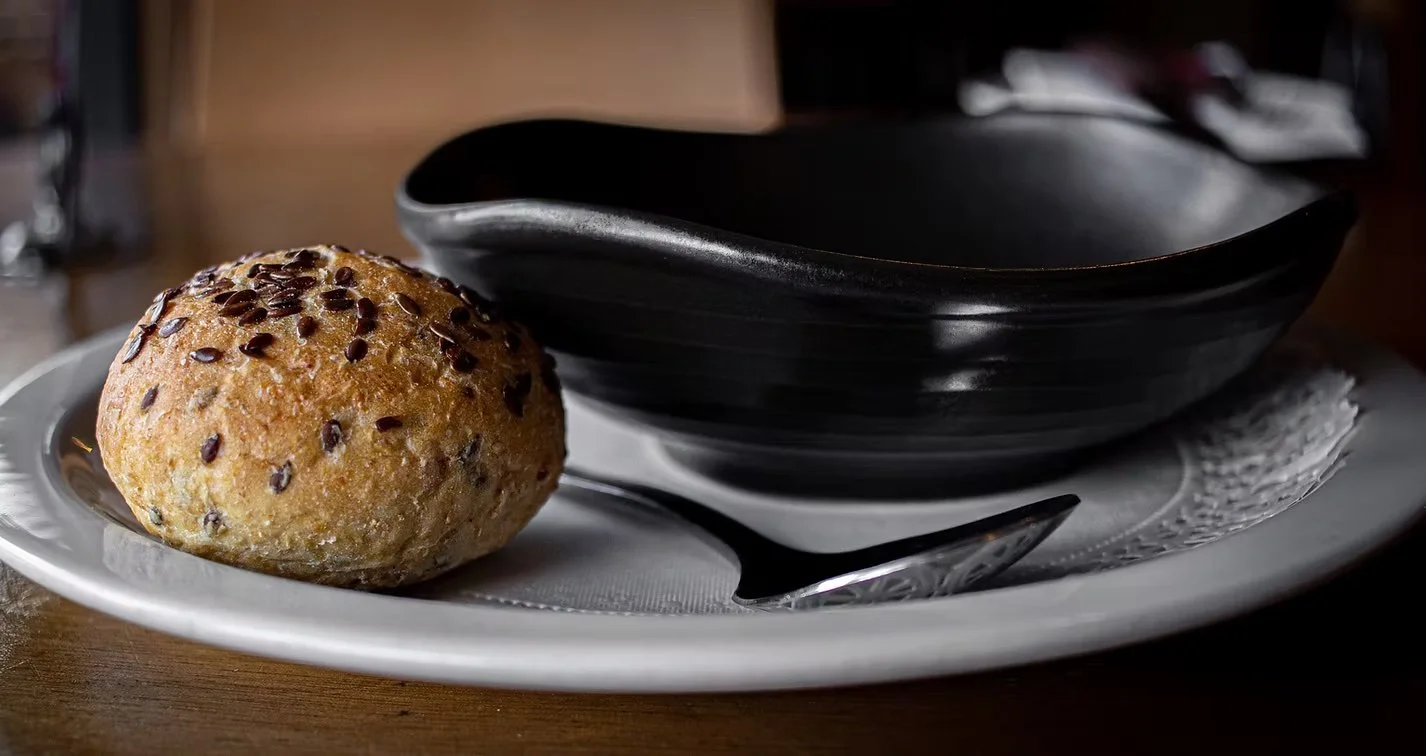 A plate with a sesame seed bun and a black bowl on a white plate, with a silver spoon beside the bowl.