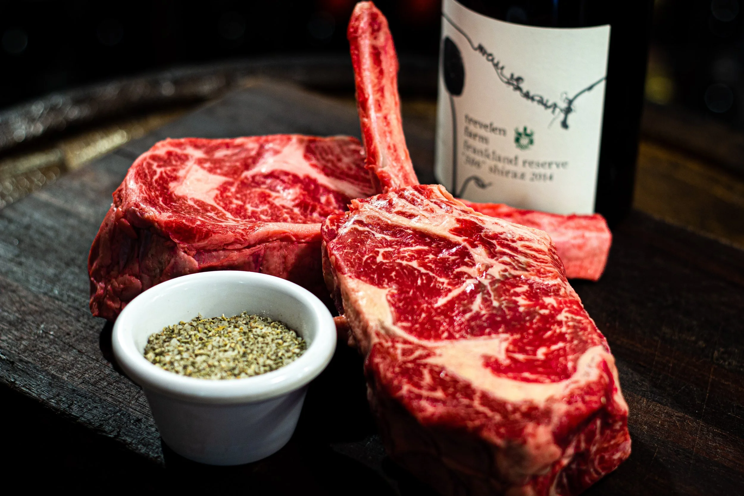 Two raw beef steaks with marbling, a small bowl of ground black pepper, and a bottle of red wine in the background, all placed on a dark wooden surface.