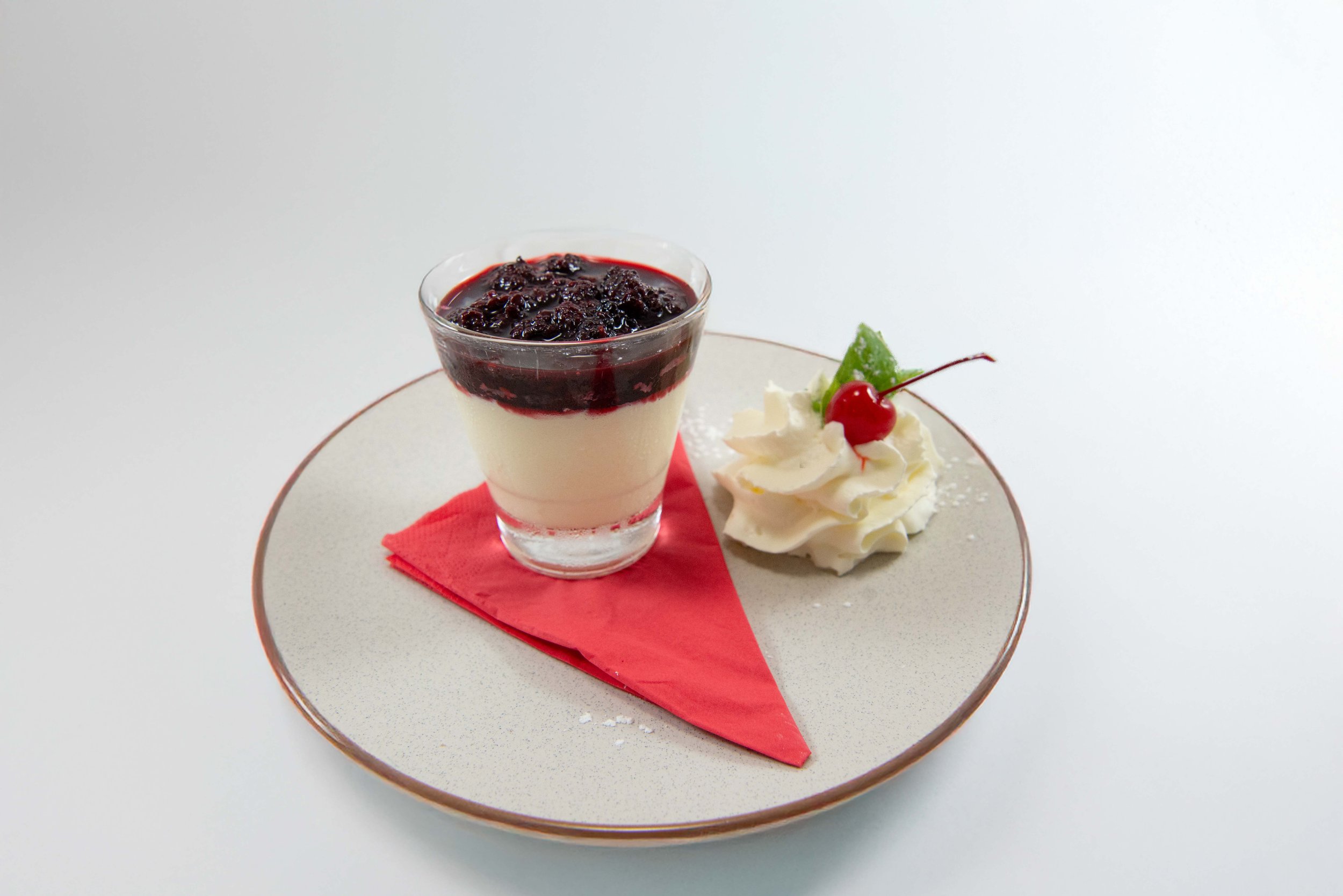 A dessert in a tall glass filled with chopped strawberries and topped with a scoop of vanilla ice cream, placed on a red plate with a red napkin on a wooden table near a window.