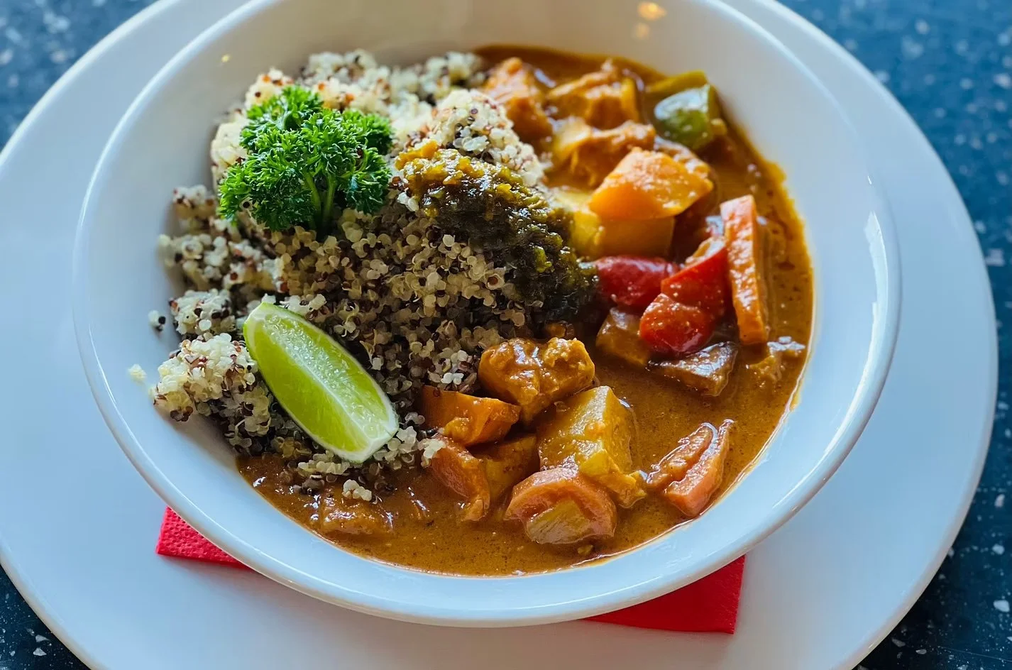 A bowl of curry with vegetables, served with quinoa, a lime wedge, and garnished with parsley on a white plate