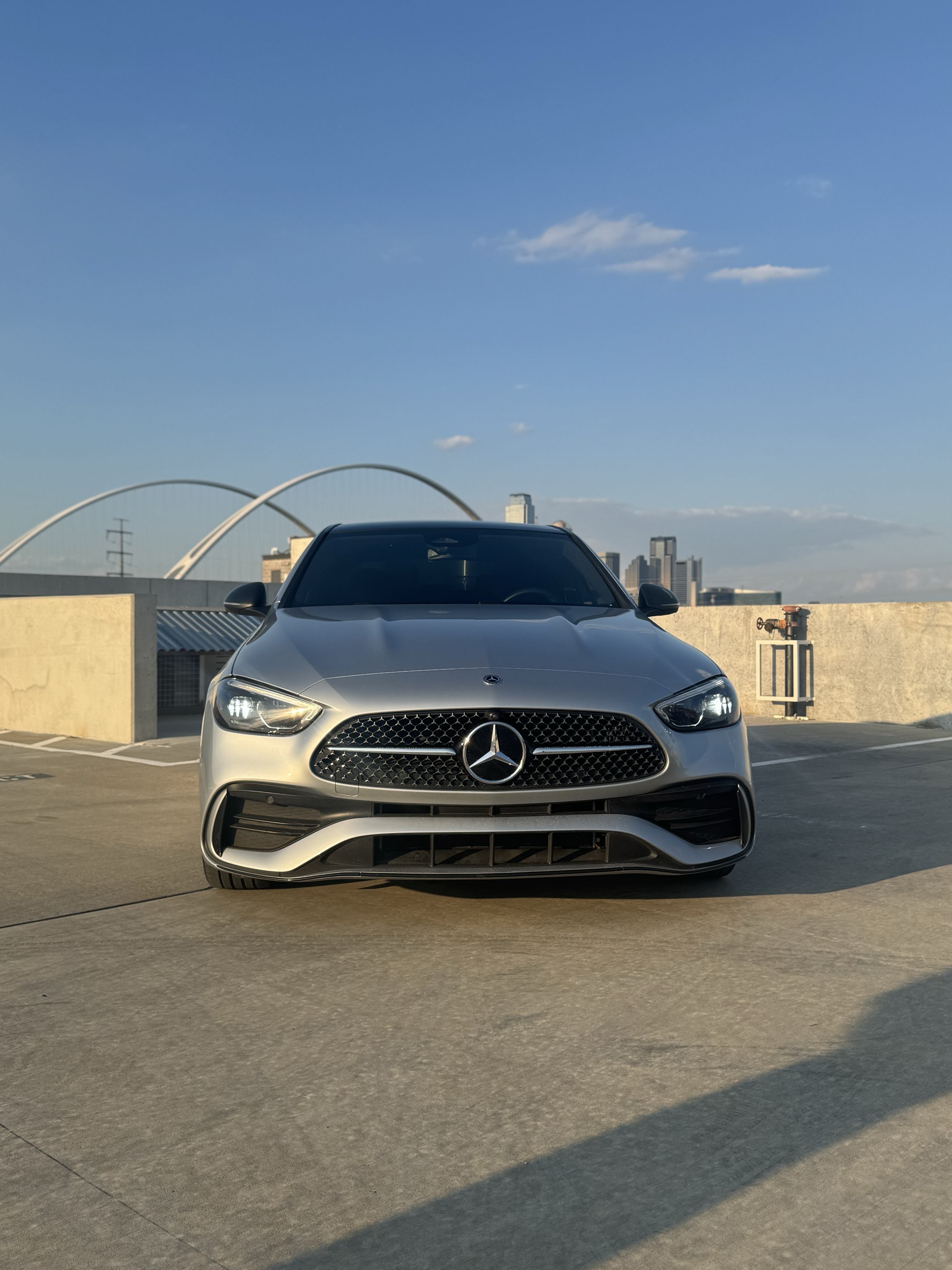 Silver Mercedes-Benz sedan parked on a rooftop parking lot with a city skyline in the background and a clear blue sky.