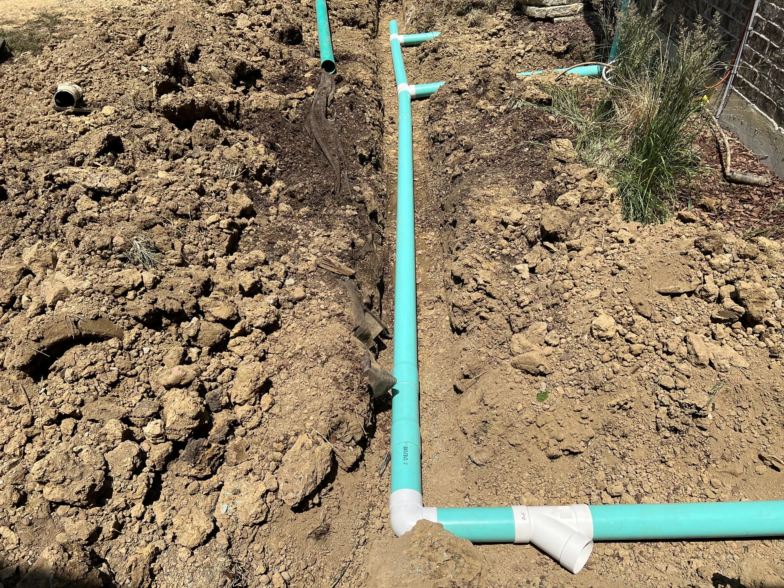 Underground plumbing pipes installed in a trench with dirt on either side and a brick wall on the right.