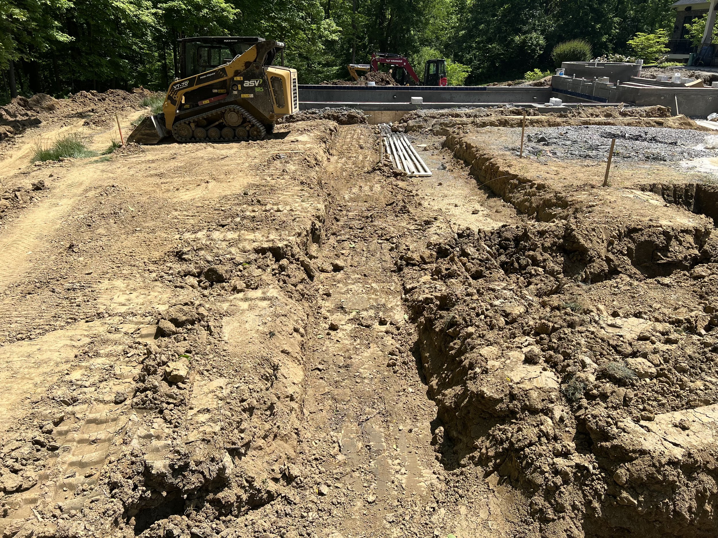 Construction site with a skid steer and excavator, trenches, and construction materials under a tree-lined sky.