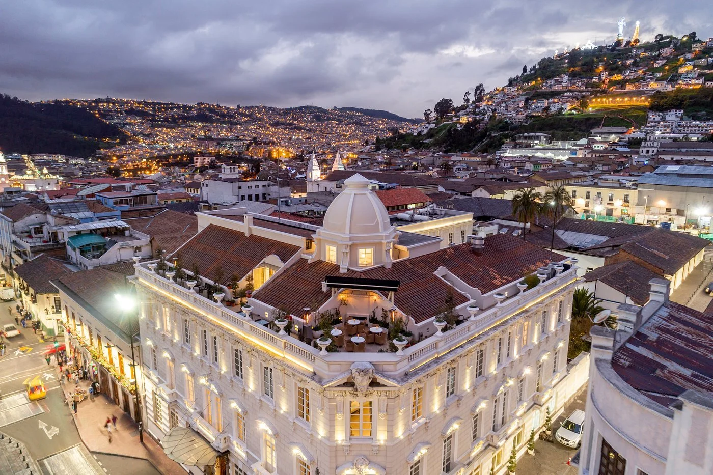 Aerial view of a cityscape at dusk with a prominent classic white building featuring a domed roof and rooftop terrace, city lights illuminating the hilly terrain with numerous houses and buildings.
