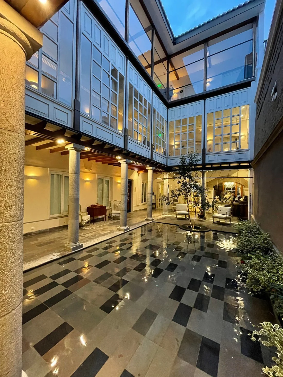 An indoor courtyard with a glossy black and white checkered tile floor, surrounded by cream-colored walls, columns, and furniture. The upper level features glass-paned windows and a glass ceiling allowing natural light to enter.