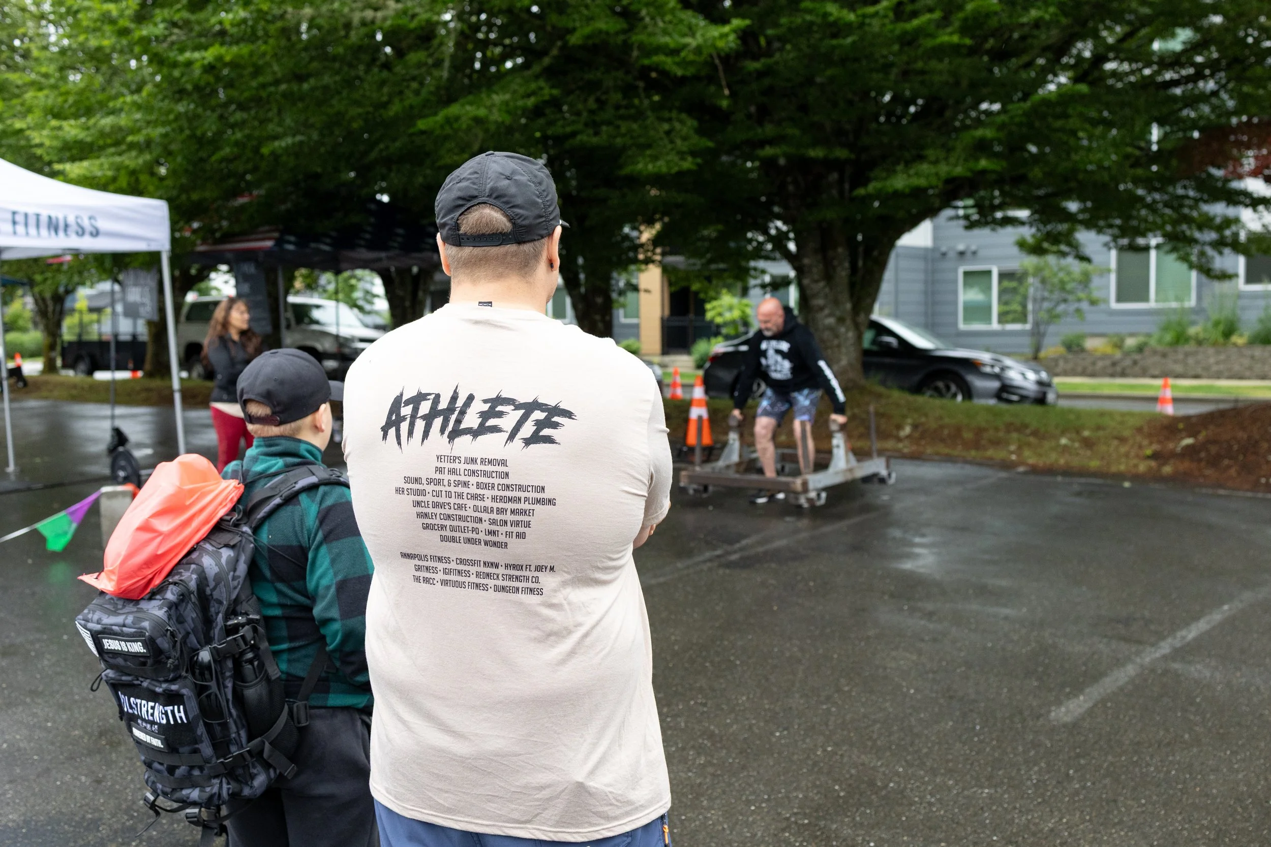 Two boys watching a man perform a fitness stunt with a platform outdoors, under a tree on a rainy day.
