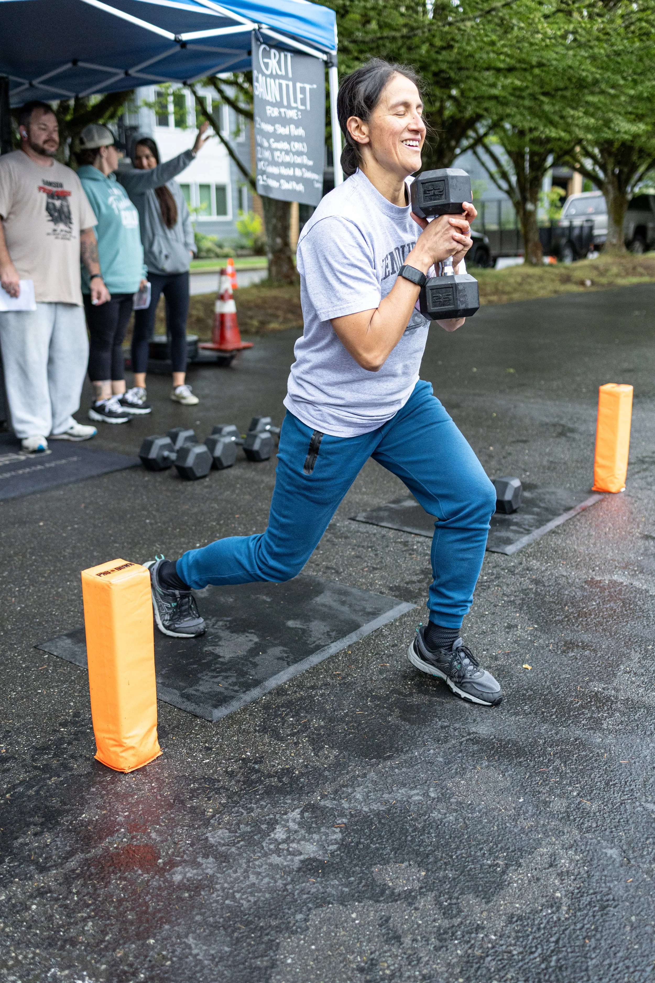 Woman performing a lunge exercise with dumbbells during outdoor fitness event, with people and sign in the background.