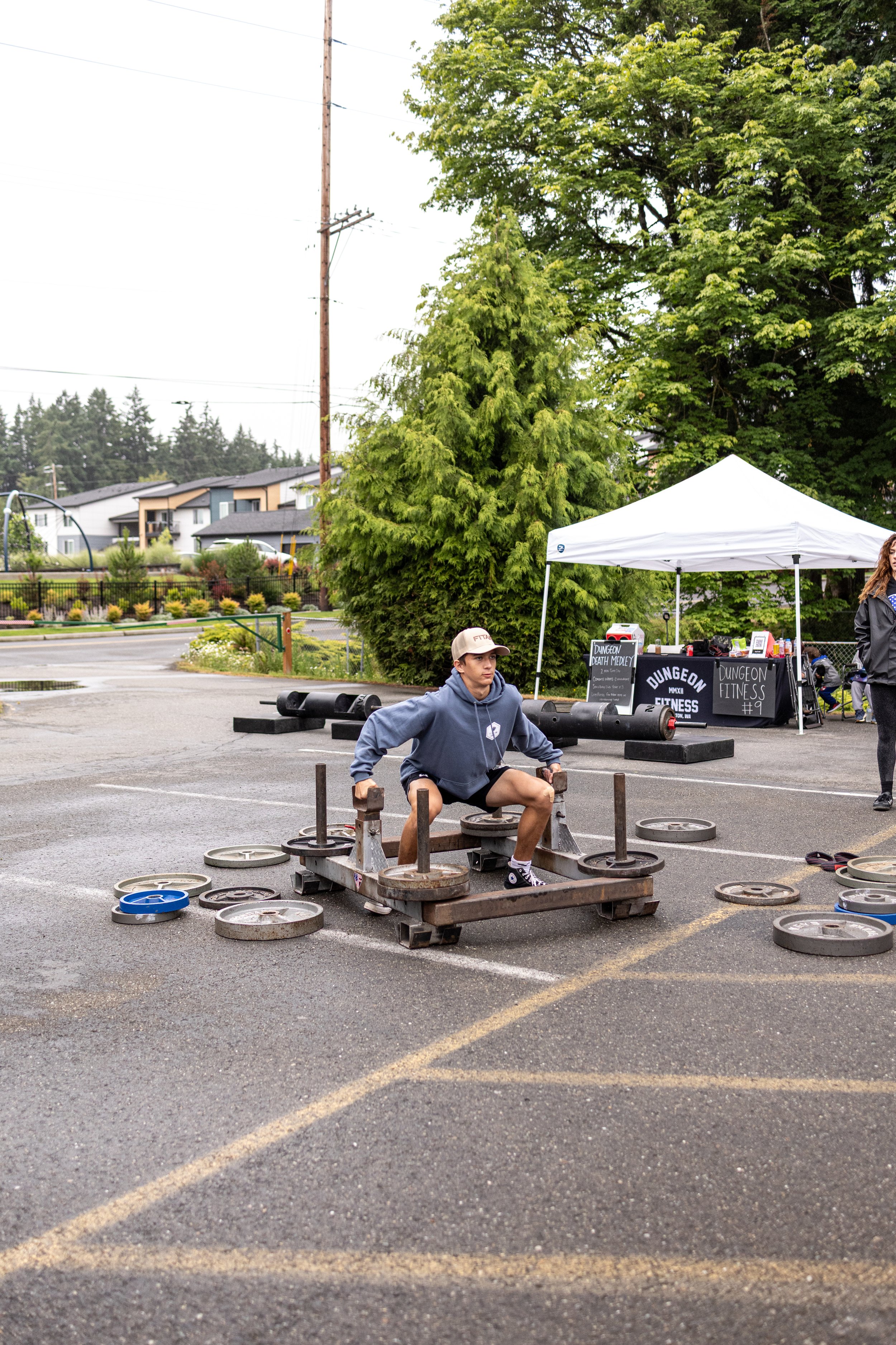 A young man in a gray hoodie and shorts lifting weights outdoors, surrounding by weight plates, at a gym event with a tent and signs in the background.