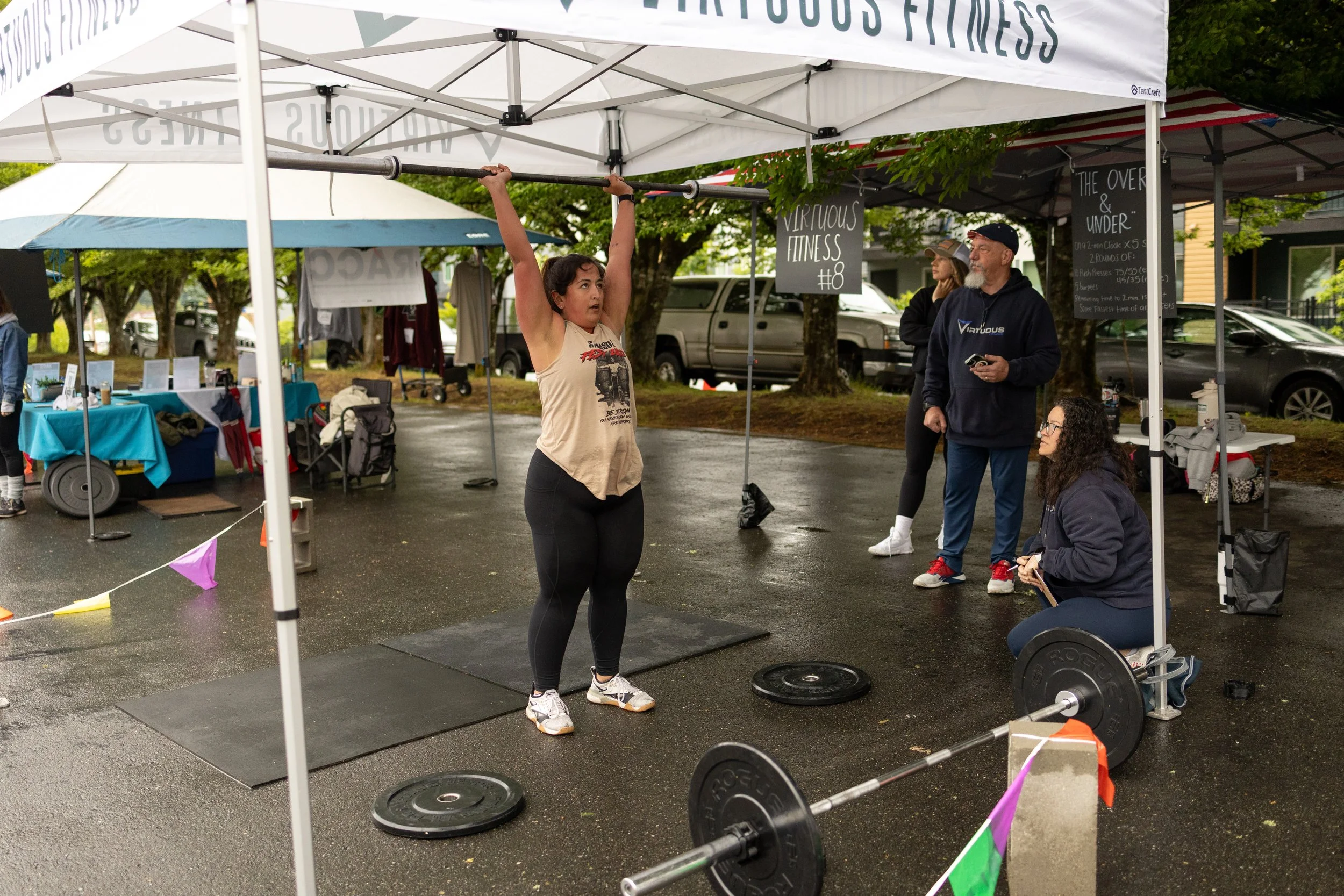 A woman is lifting a barbell overhead at an outdoor fitness event under a white tent on a wet pavement. There are weights on the ground and a barbell on a stand, with a barbell and weights in the foreground. Several people are observing or participat
