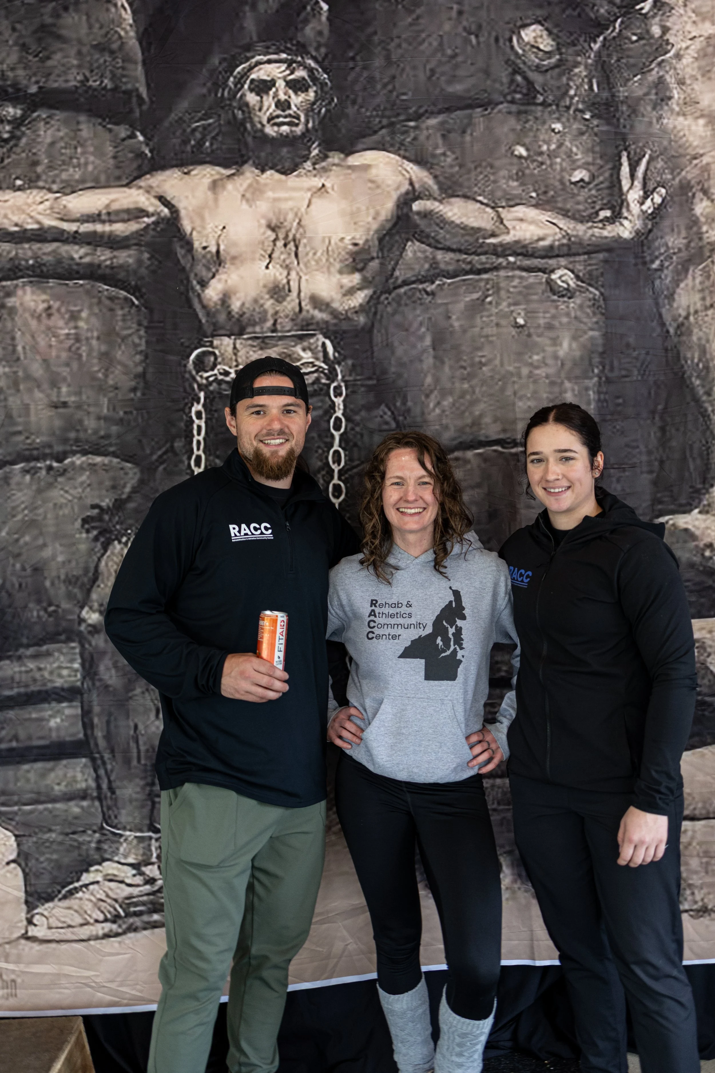 Three people standing in front of a large mural of Jesus Christ on the cross. The man on the left is holding a can of drink, and all three are smiling. The woman in the middle is wearing a gray hoodie with a map of New York and text. The woman on the